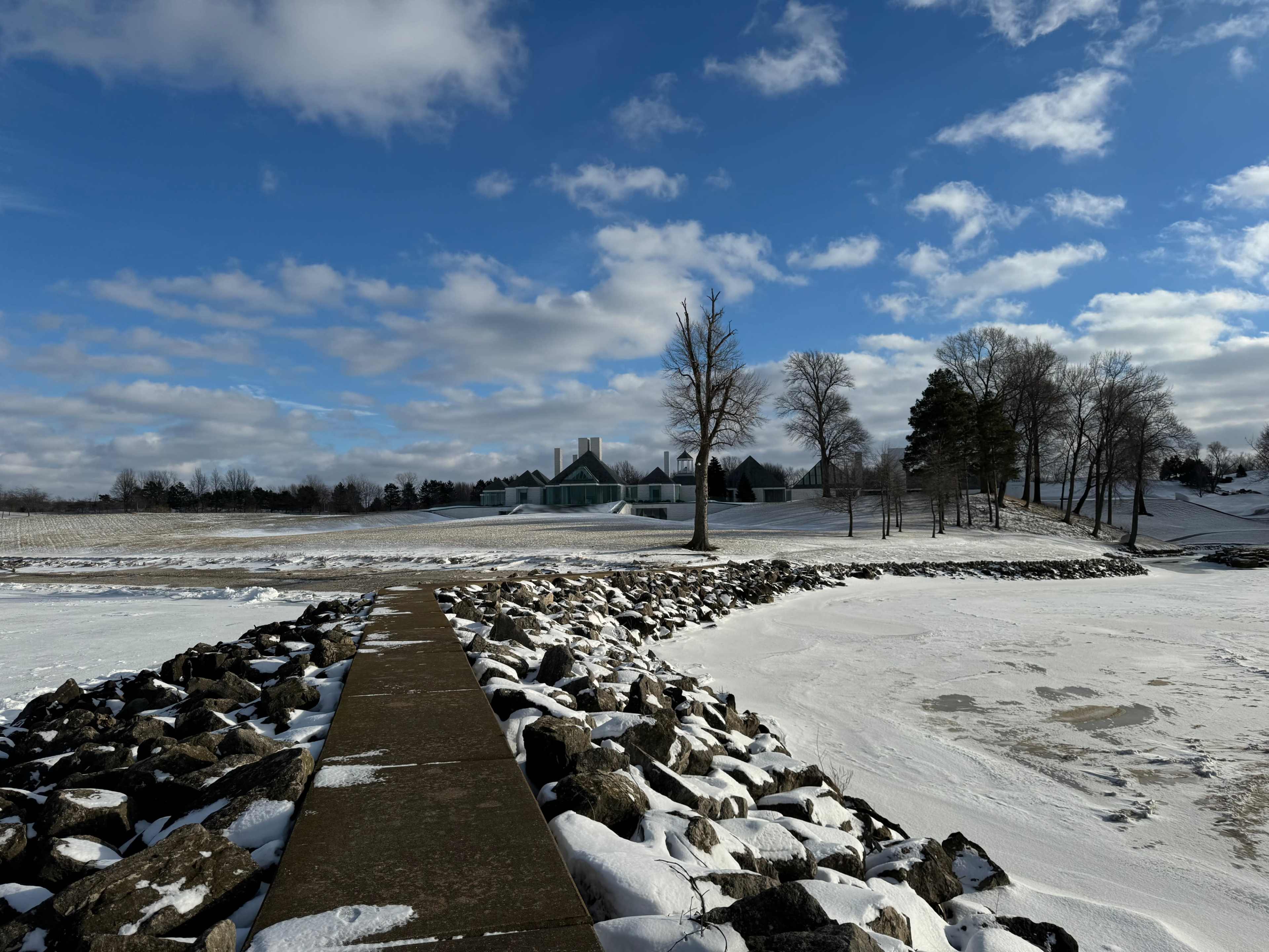 A paved walkway leads past a stone barrier along a snowy landscape, with a large building and bare trees in the background under a partly cloudy sky.