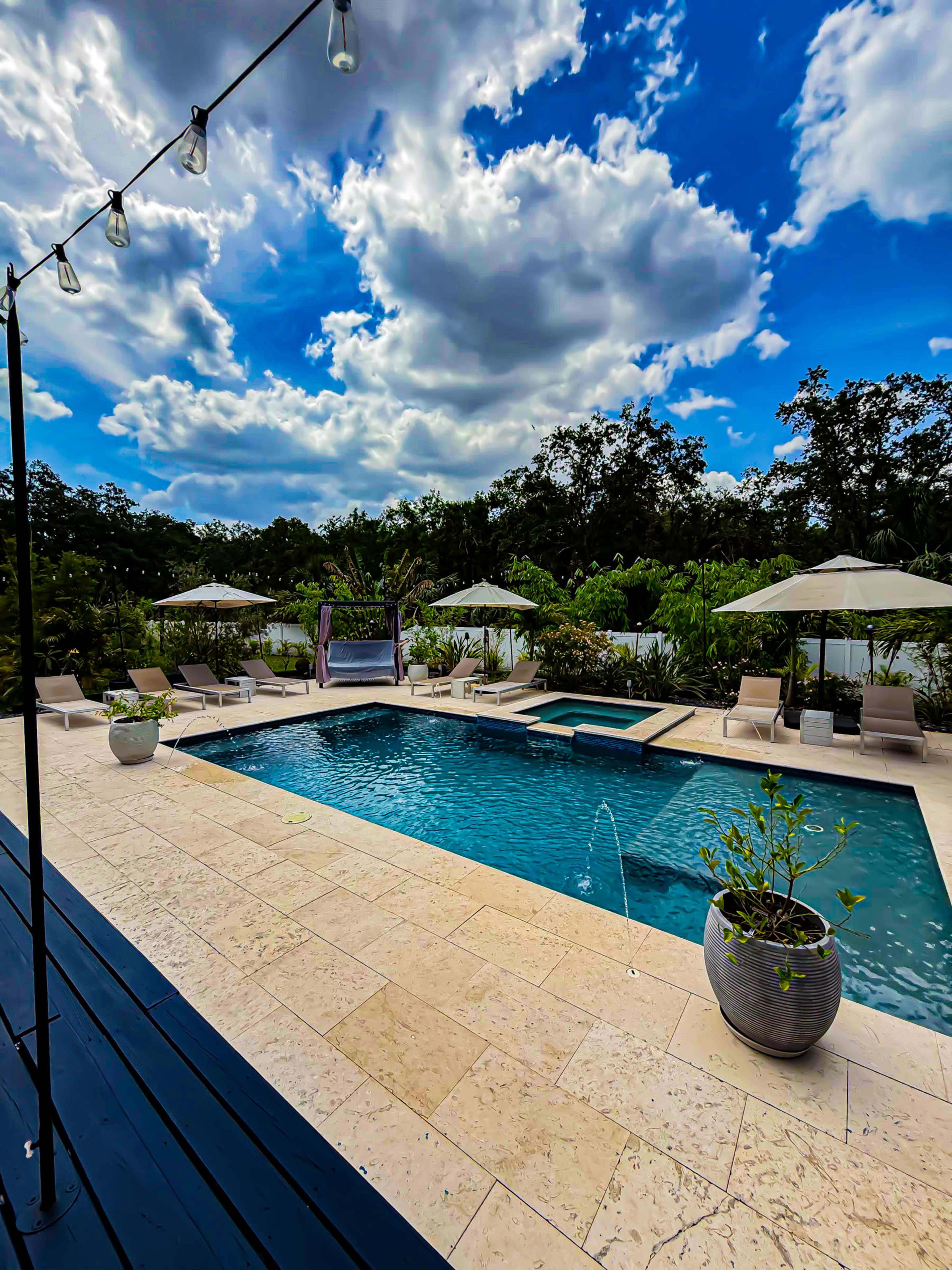 The image features a landscaped outdoor pool area surrounded by lounge chairs and umbrellas under a blue sky with clouds.