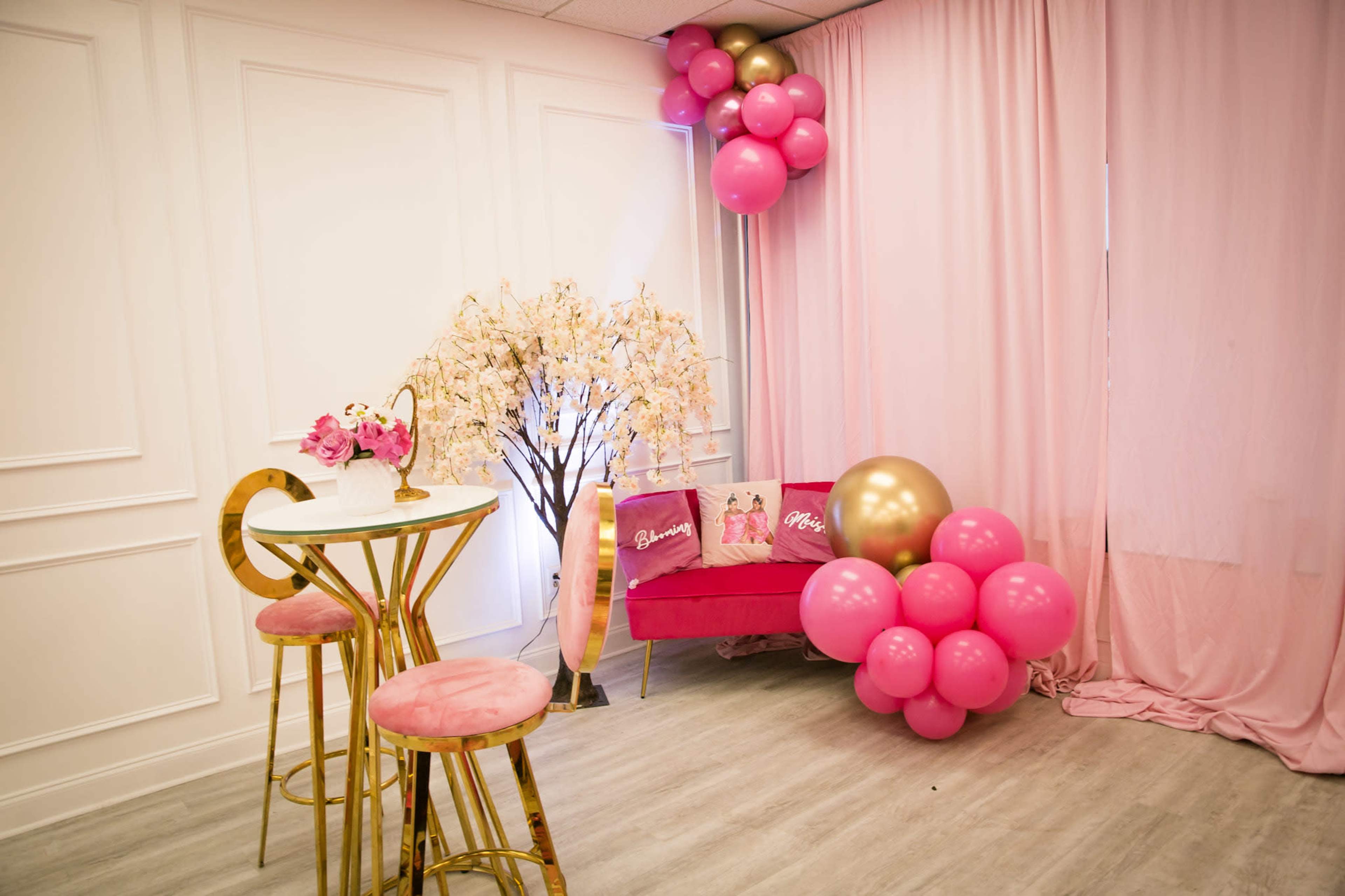 A pink-themed room with a small table and two chairs, a decorative flower tree, and a pink couch adorned with balloons.