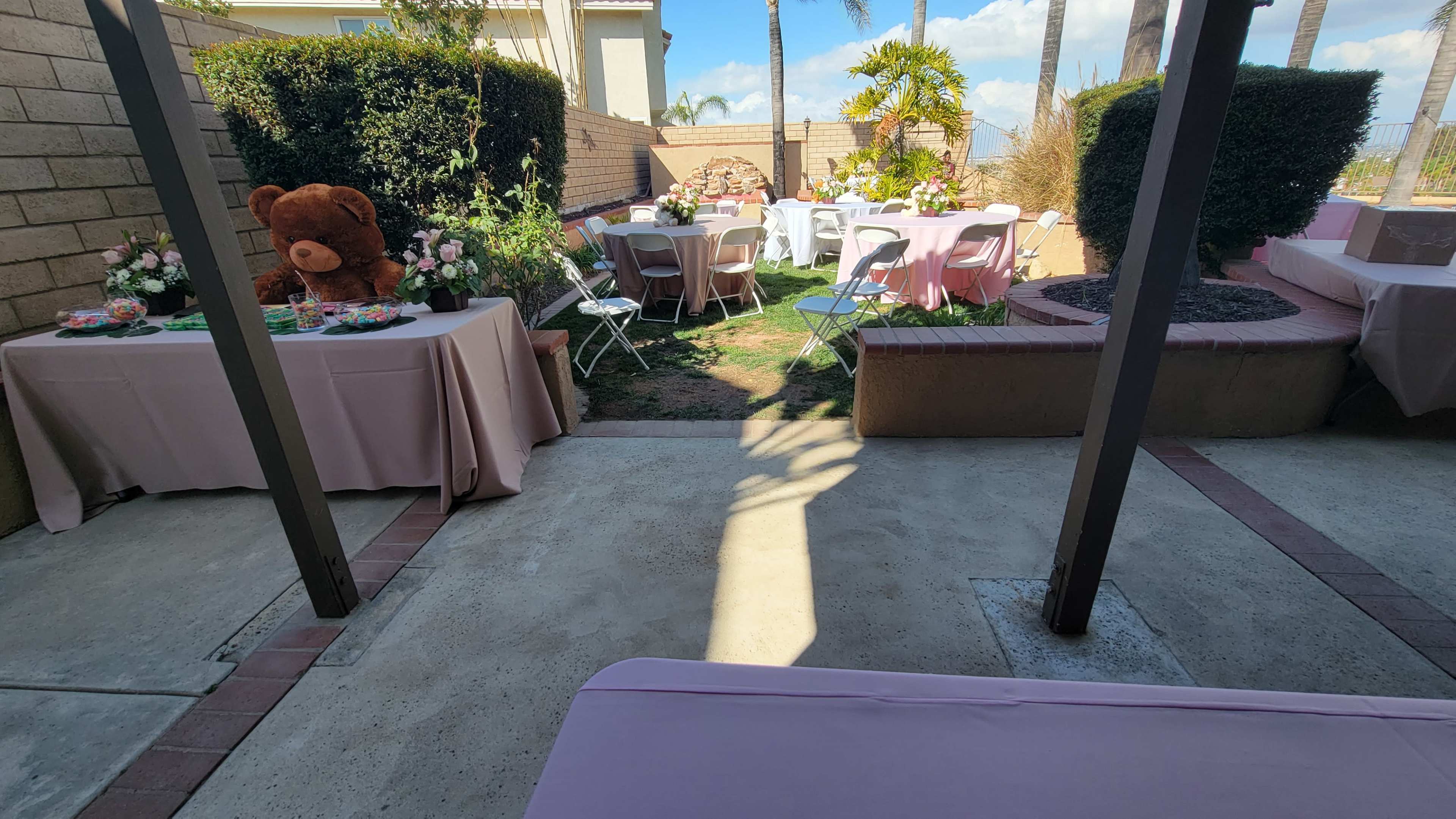 The scene shows an outdoor patio set up for a gathering, featuring tables covered with pink tablecloths, a large teddy bear, floral centerpieces, and a lawn area with palm trees in the background.