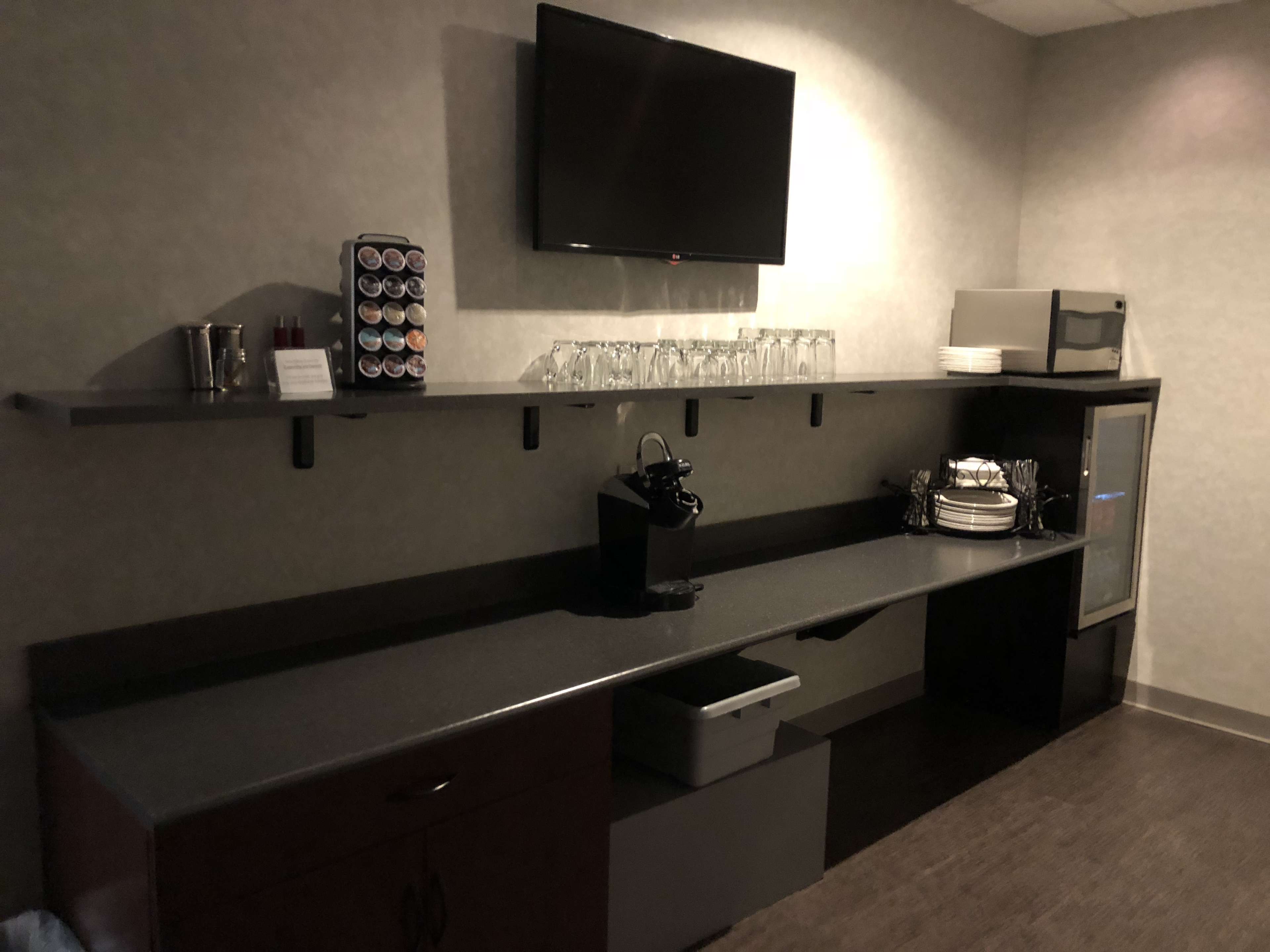 A modern kitchenette area features a countertop with a coffee machine, cups, a microwave, and a small refrigerator beneath a wall-mounted television.
