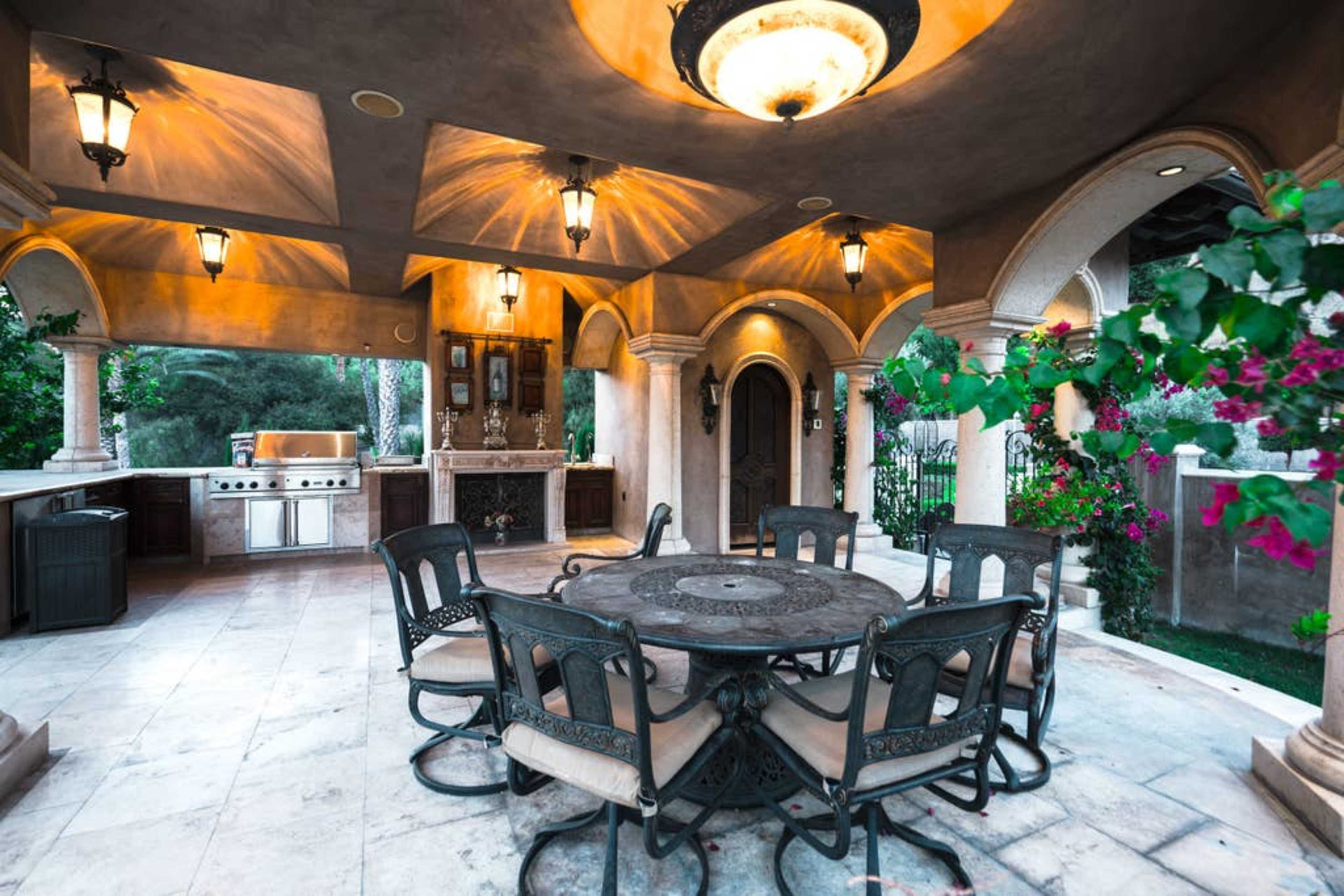 The image shows a spacious outdoor dining area with a round table and wrought-iron chairs, surrounded by decorative stone columns and an outdoor kitchen.