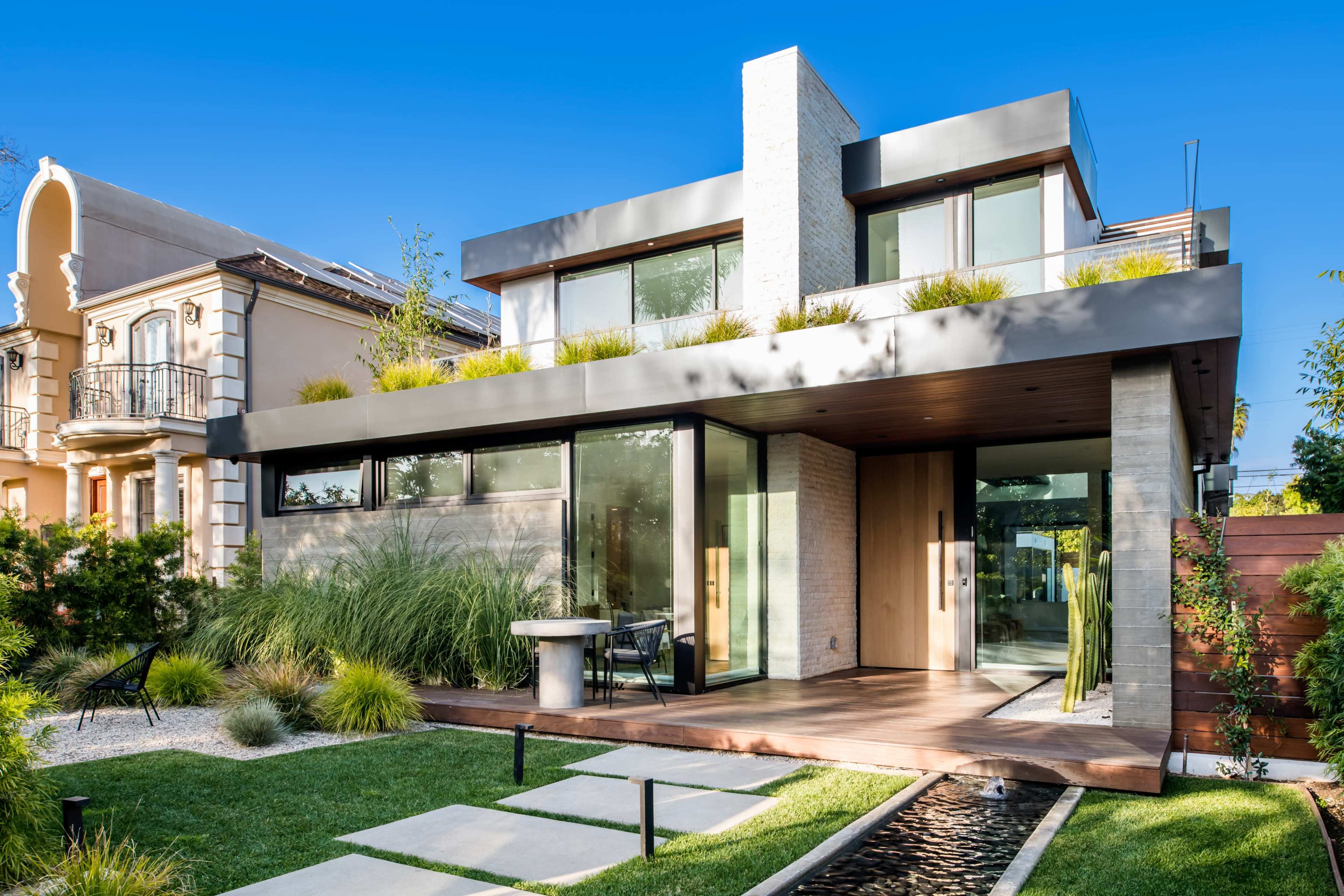 A modern two-story house with expansive glass windows, a flat roof, and a landscaped front yard with stone pathways and greenery.