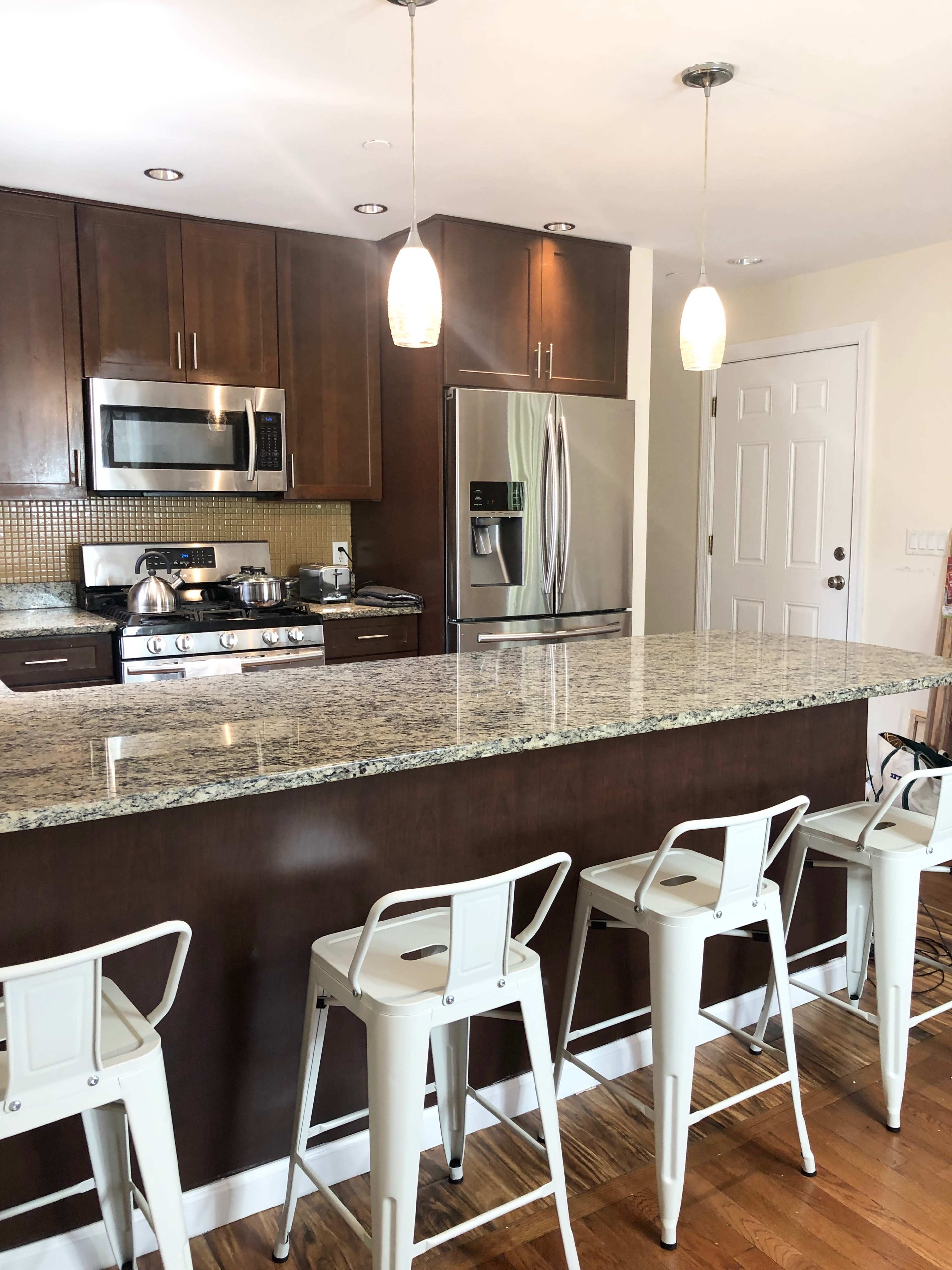 A modern kitchen features dark wood cabinetry, stainless steel appliances, and a granite countertop island with white bar stools.