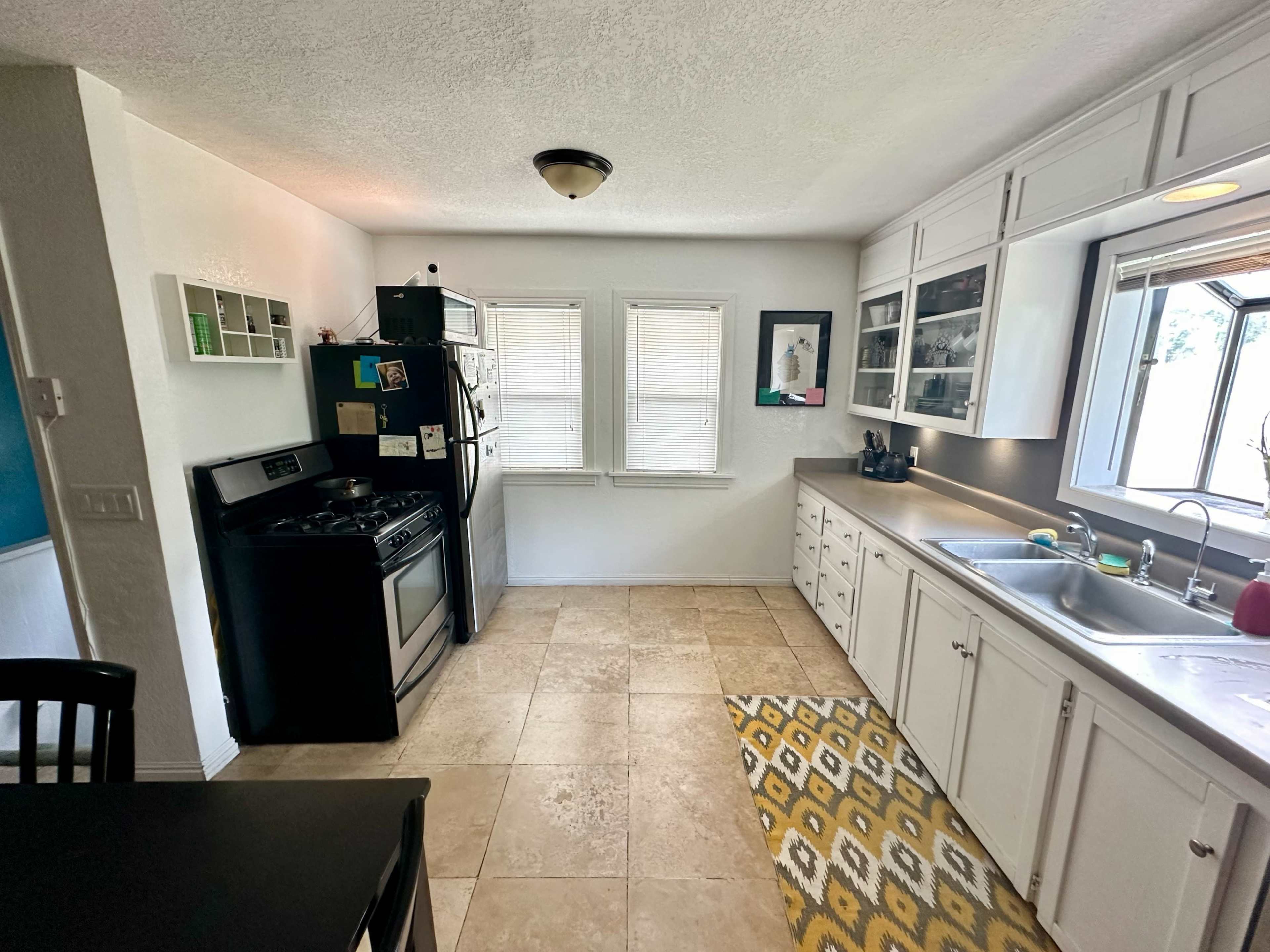 A kitchen with light-colored cabinets, a black stove, and a sink beside a window, featuring tiled flooring and a patterned rug.