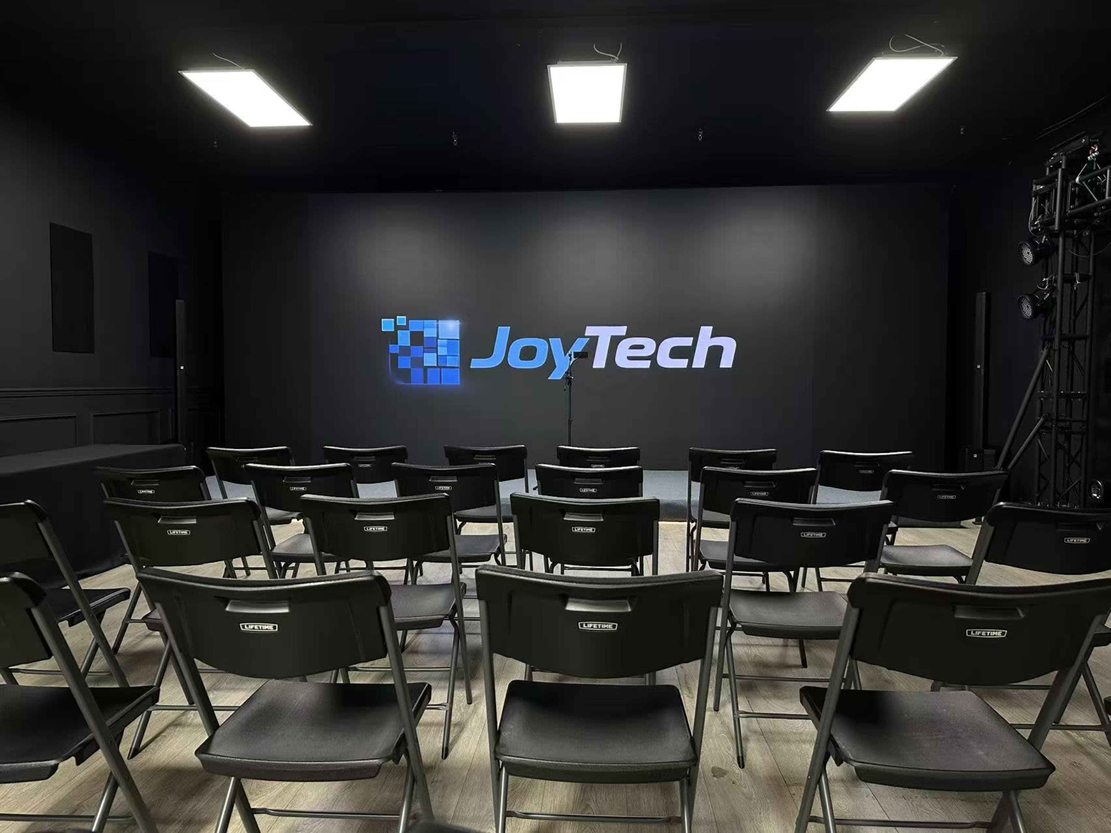 The image shows a minimalist presentation room with rows of black folding chairs facing a large screen displaying the logo "JoyTech" on a dark wall.