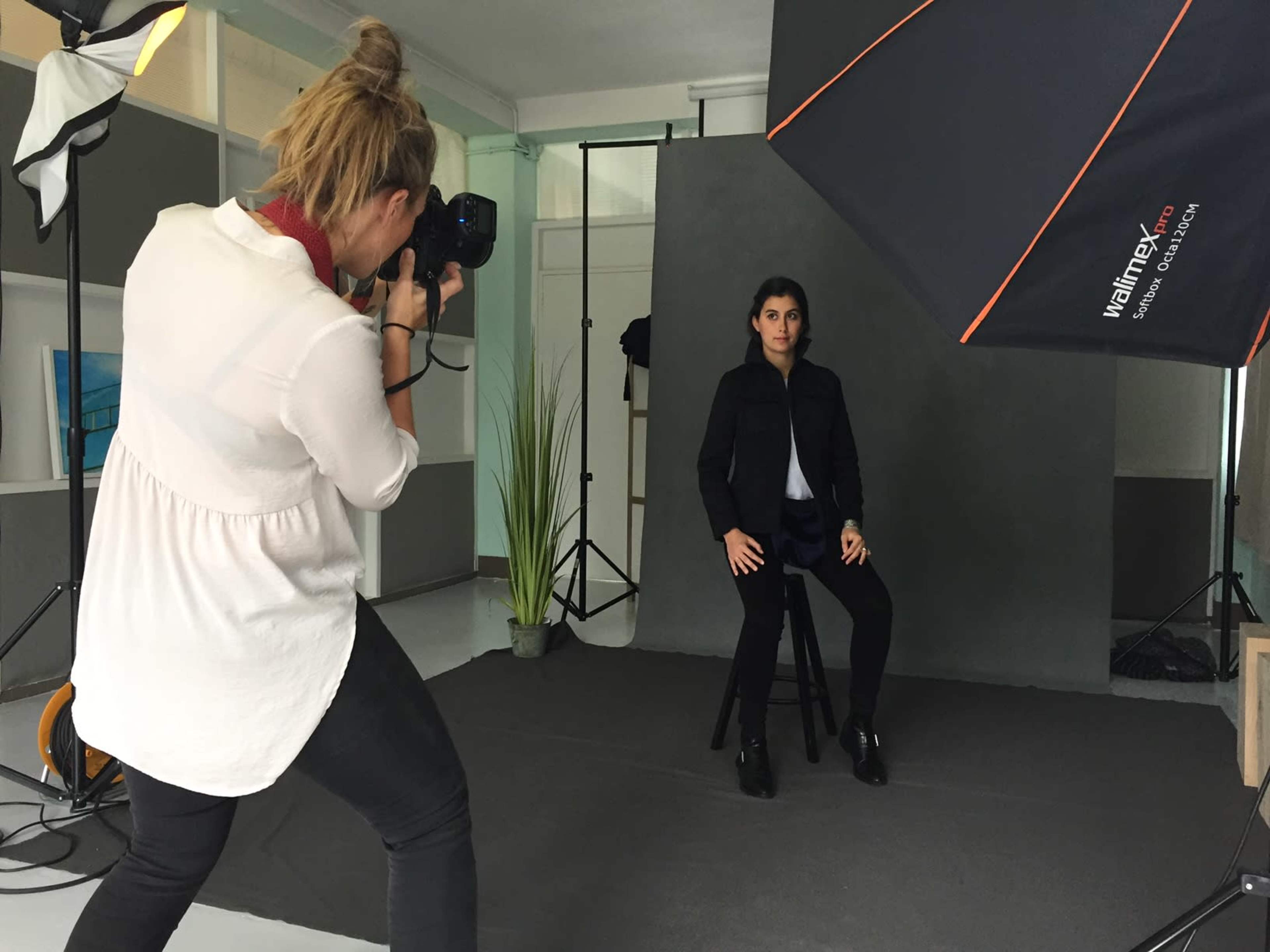 A photographer is capturing images of a seated model in a studio setting, surrounded by lighting equipment and a backdrop.