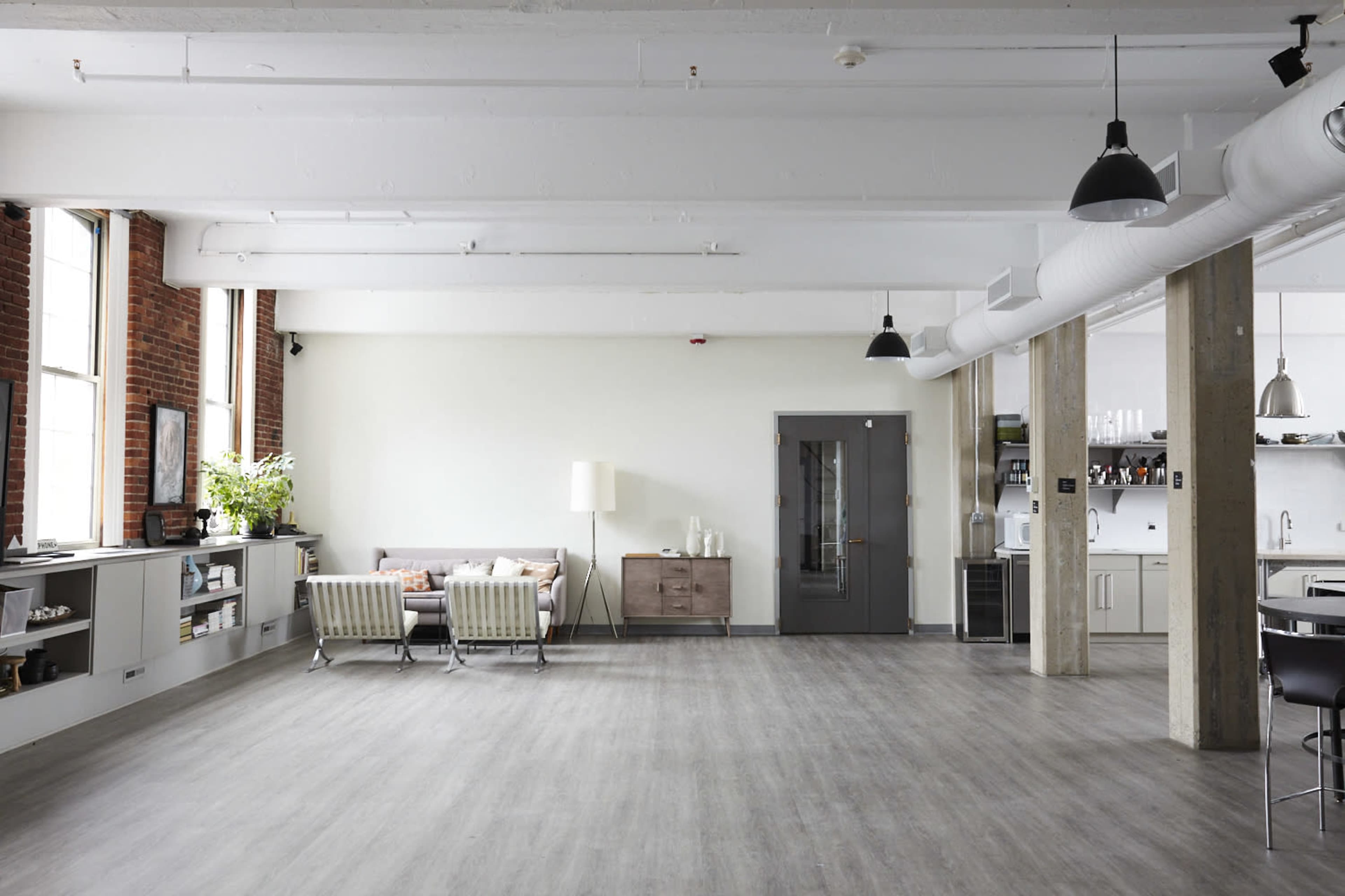 A spacious, minimalistic loft features large windows, a modern sofa, a wooden cabinet, and an open kitchen area with a dining table.