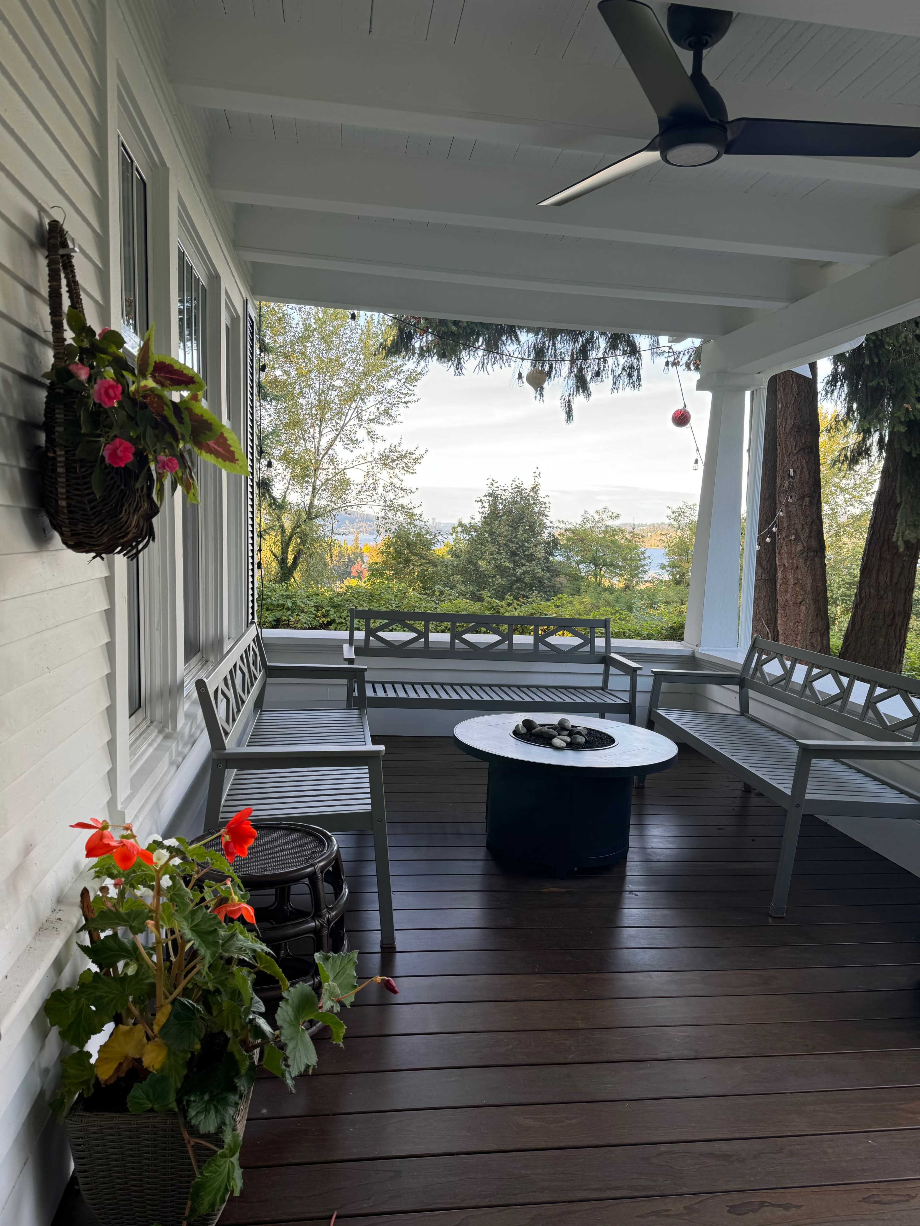 The image shows a porch with gray benches and a round table, surrounded by greenery and a scenic view in the background.