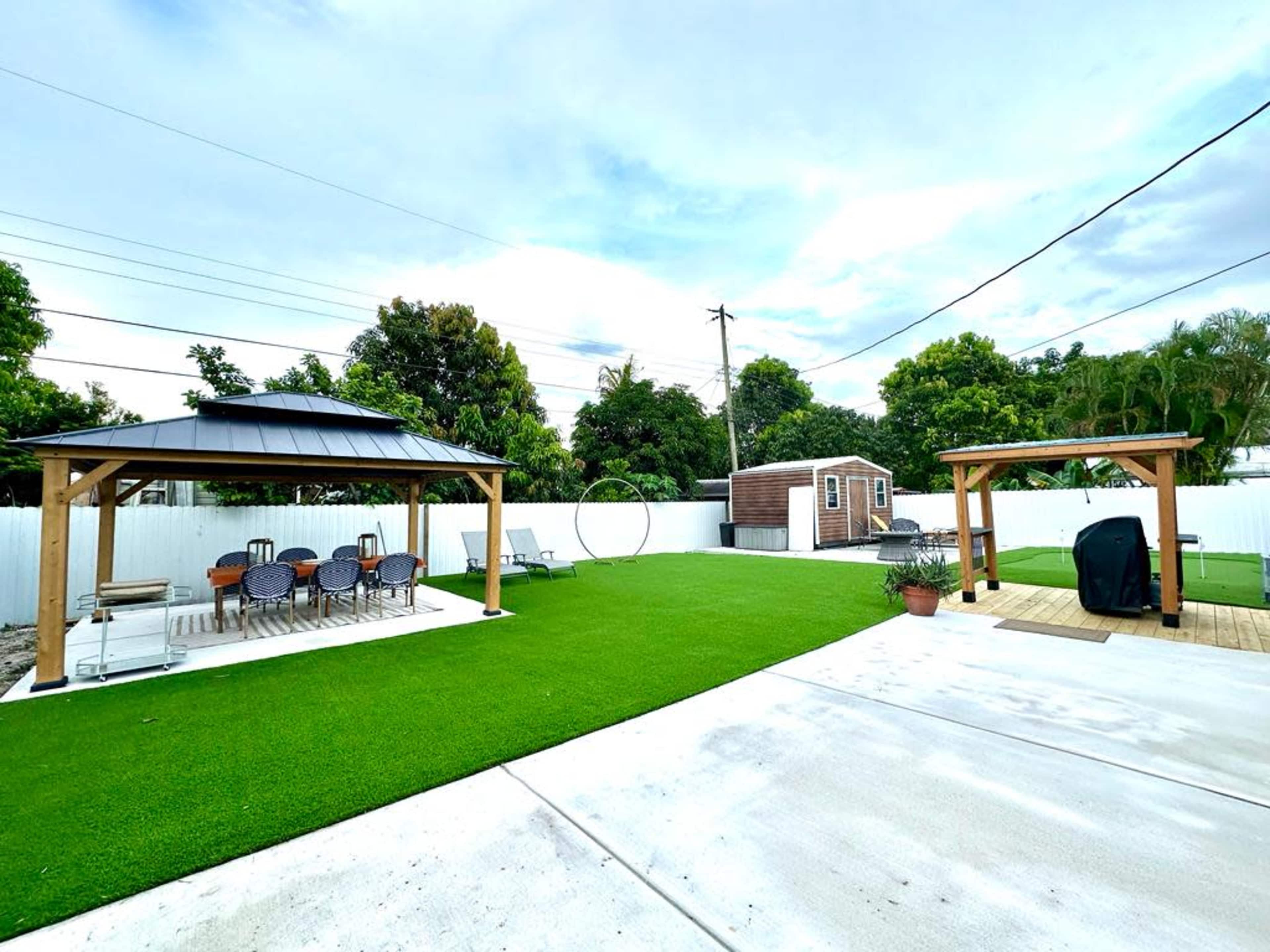 The image shows a landscaped backyard featuring two wooden gazebos, a patch of artificial turf, and a storage shed in a fenced area.