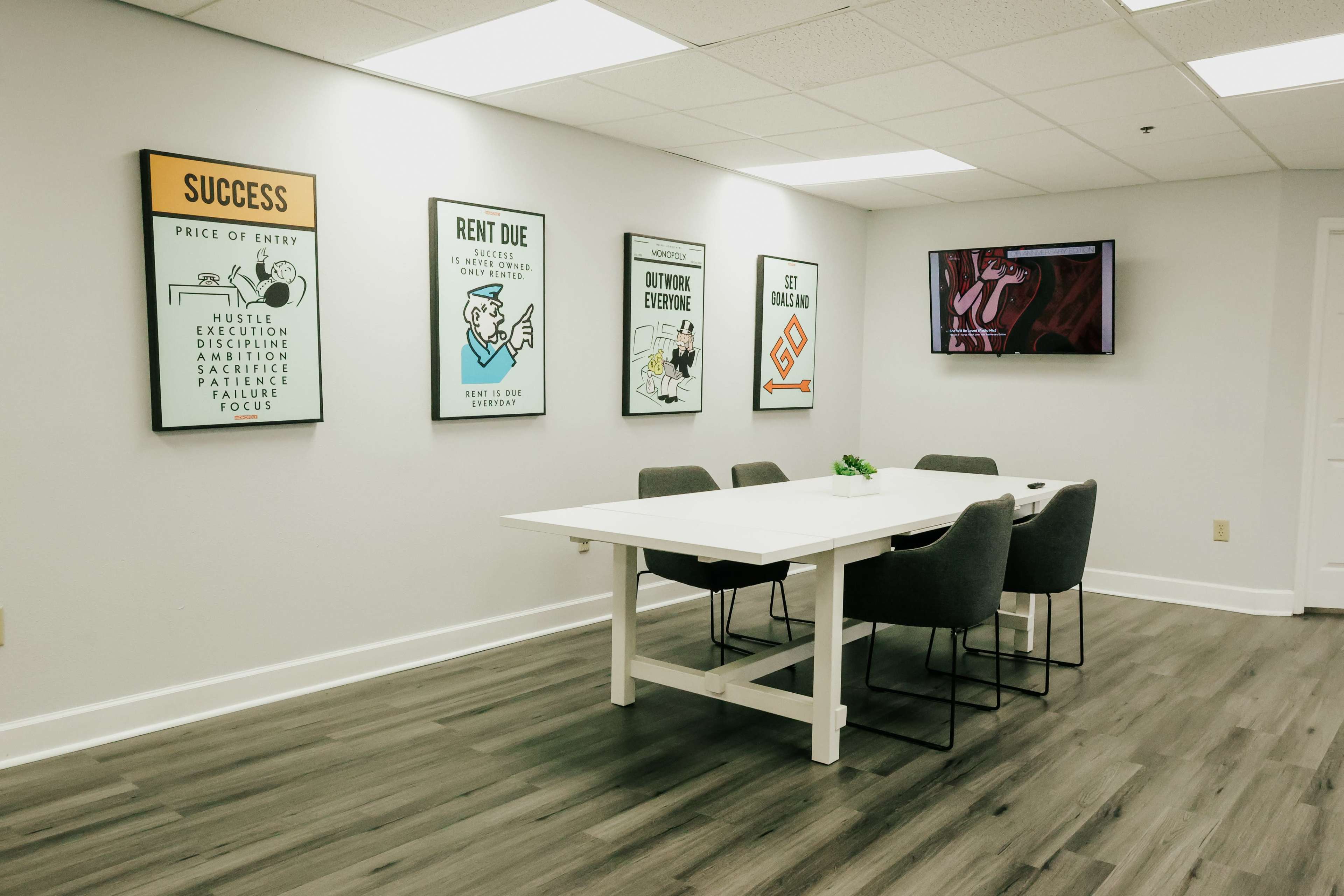 A modern meeting room features a white table surrounded by gray chairs, with framed motivational posters on the wall.