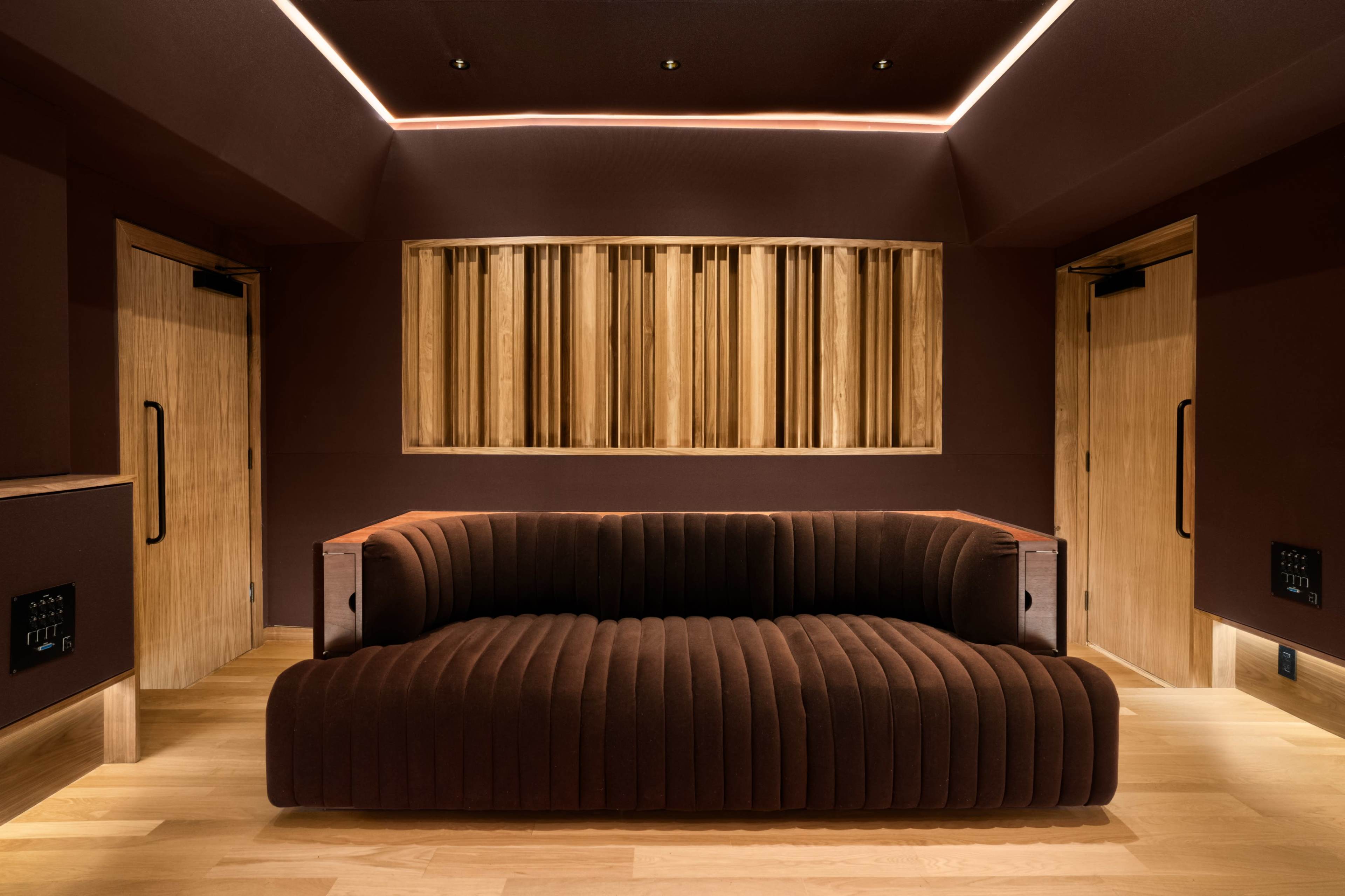 The image depicts a modern lounge area featuring a plush brown sofa against a backdrop of wooden paneling and two wooden doors.