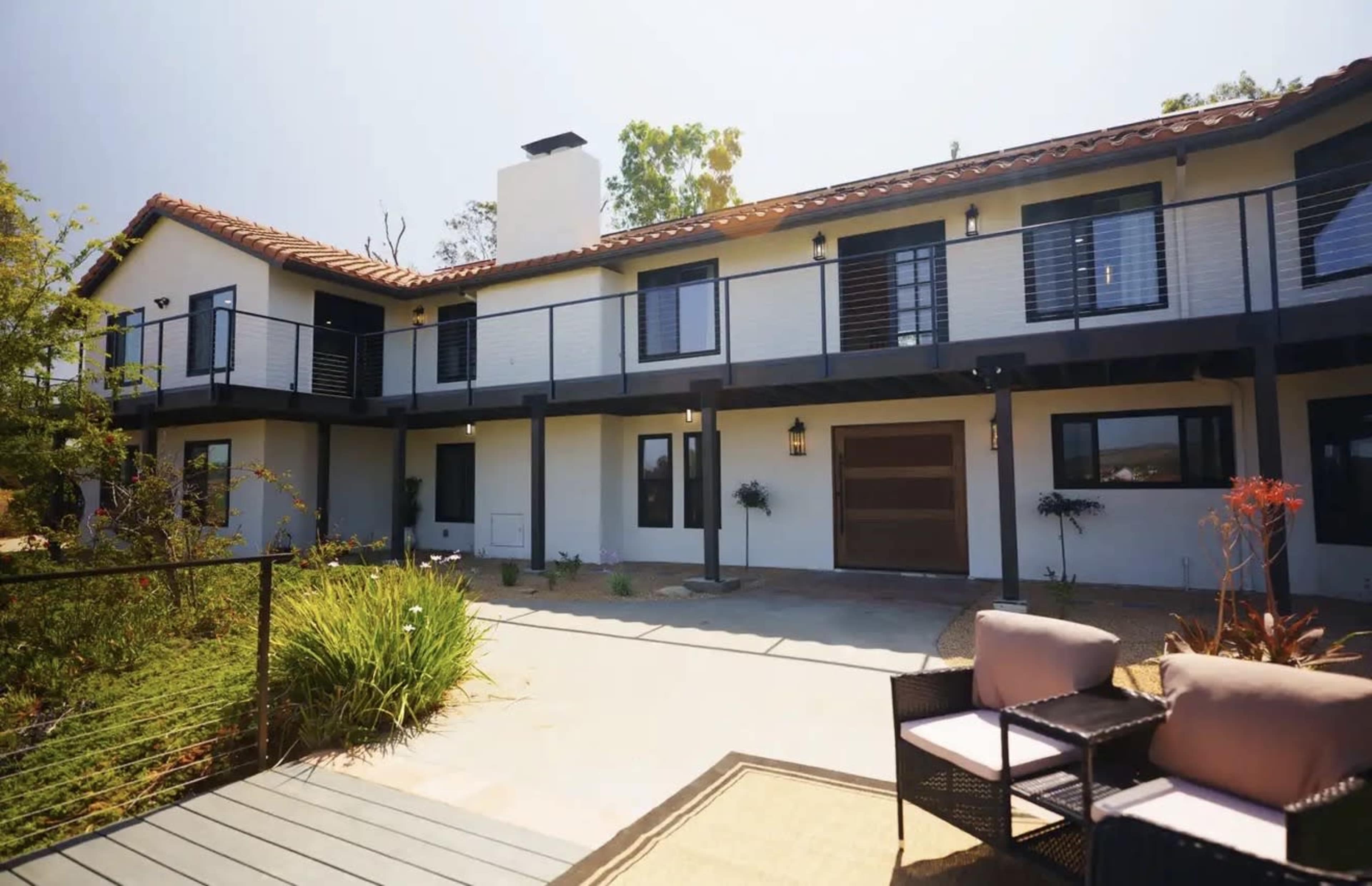 The image shows a modern two-story house with a balcony, surrounded by a landscaped patio area featuring outdoor seating.