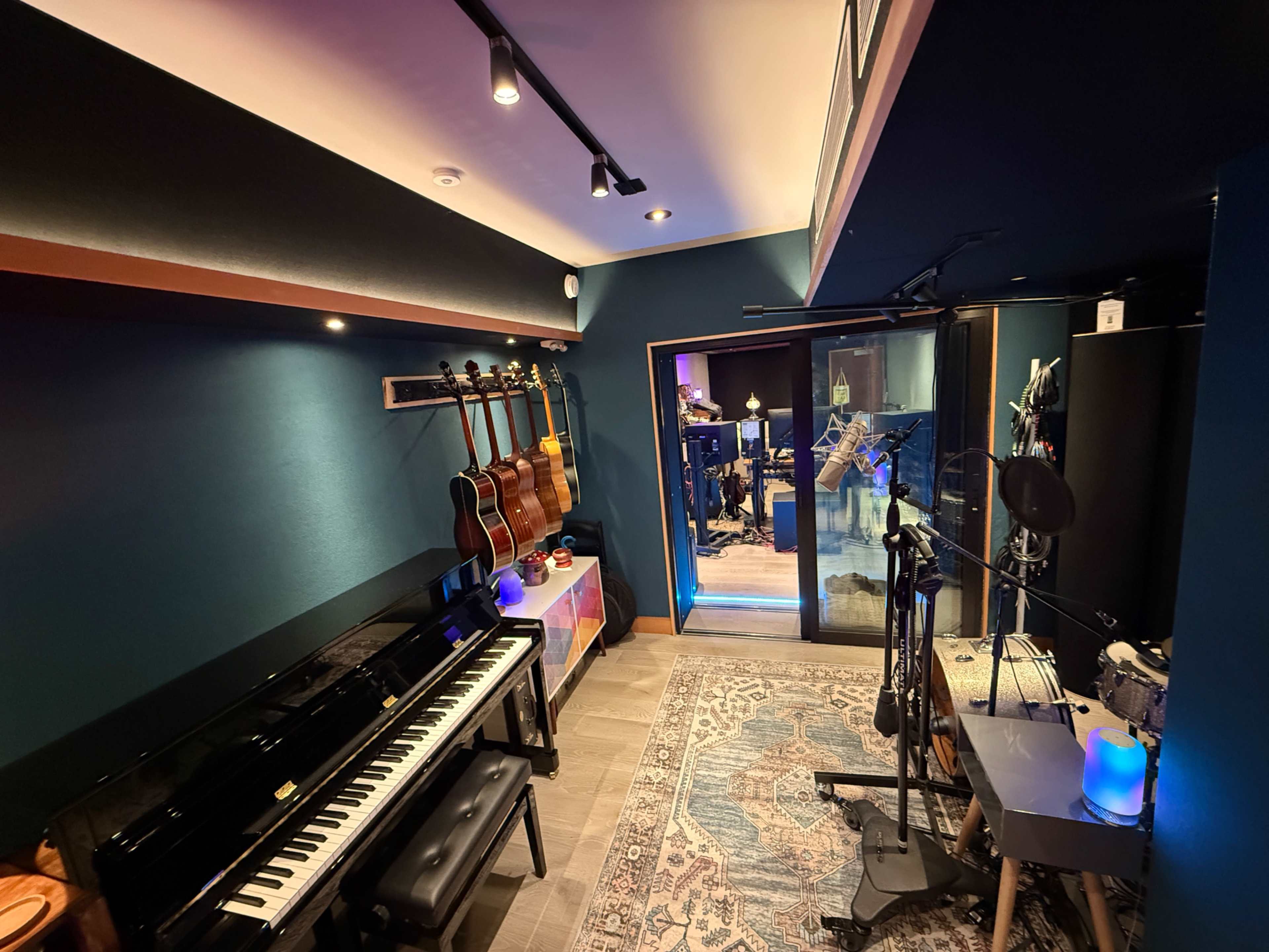 The image shows a recording studio featuring a piano, several guitars hanging on the wall, and various microphones set up in a dimly lit, colorful environment.