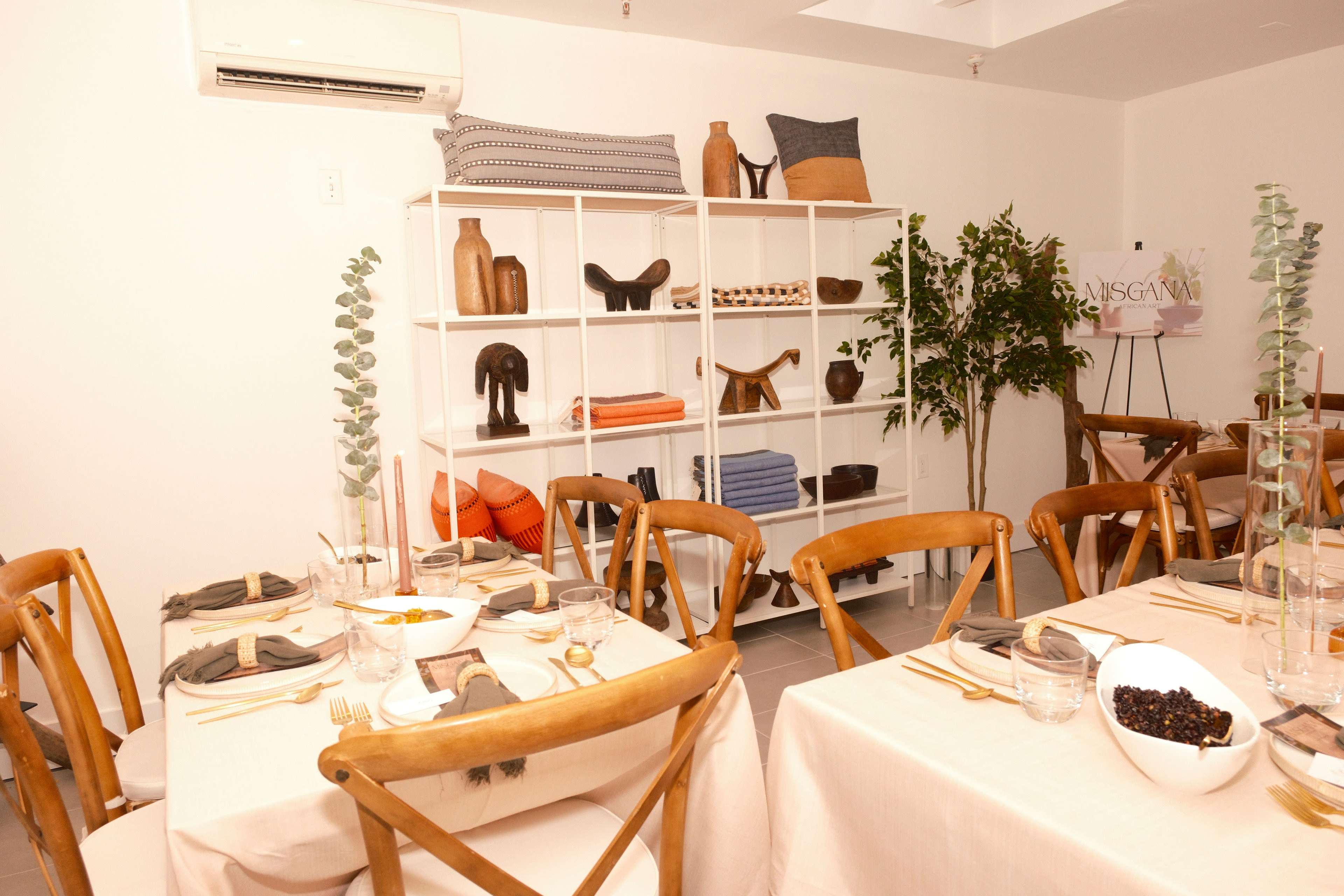 The image shows a modern dining setup with wooden chairs, a table set with dishes, and shelves displaying various decor items in a light-colored room.