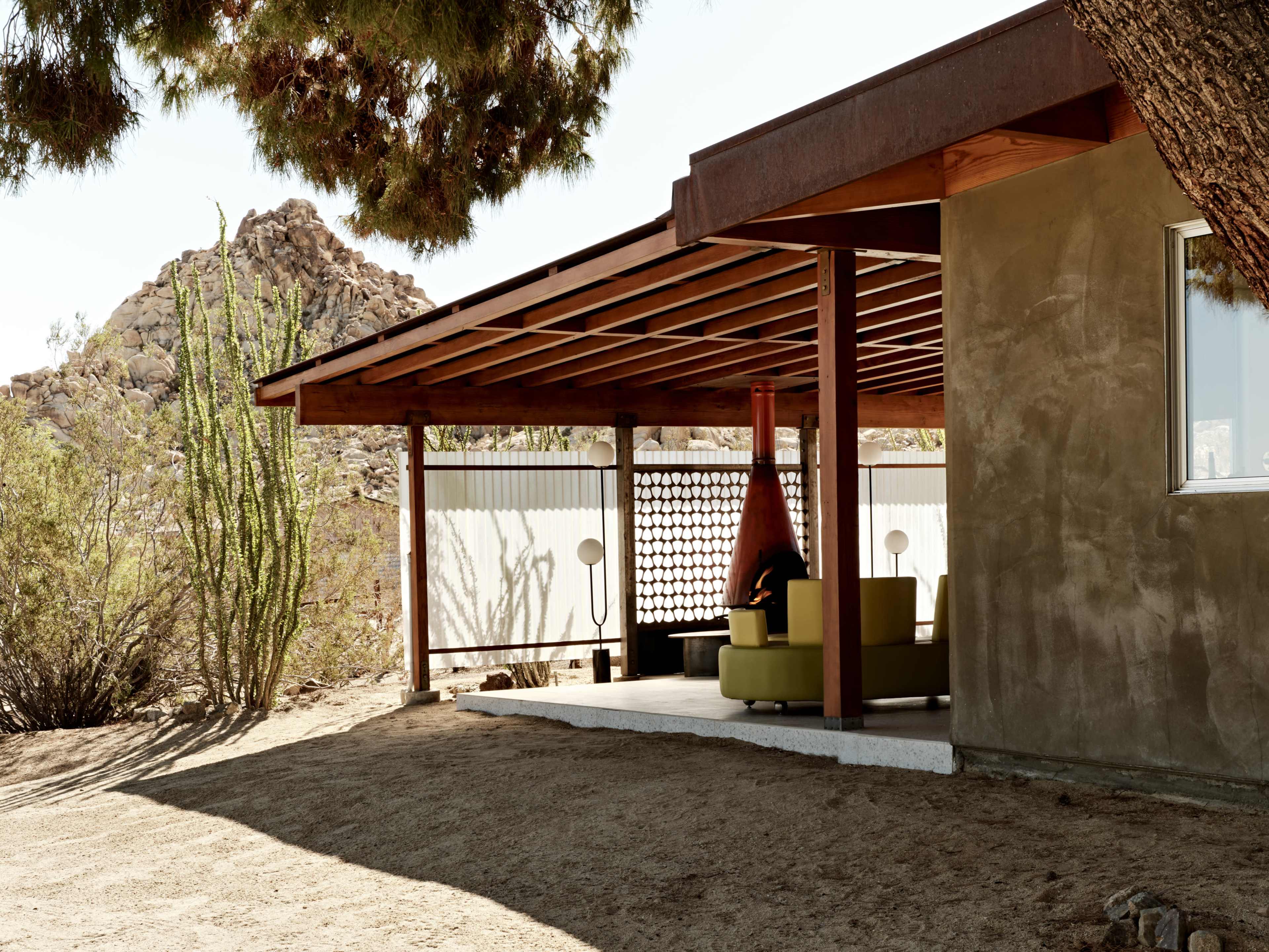 The image shows a modern, open-air living space with a green sofa and a distinctive red fireplace, set against a backdrop of rocky terrain and sparse vegetation.