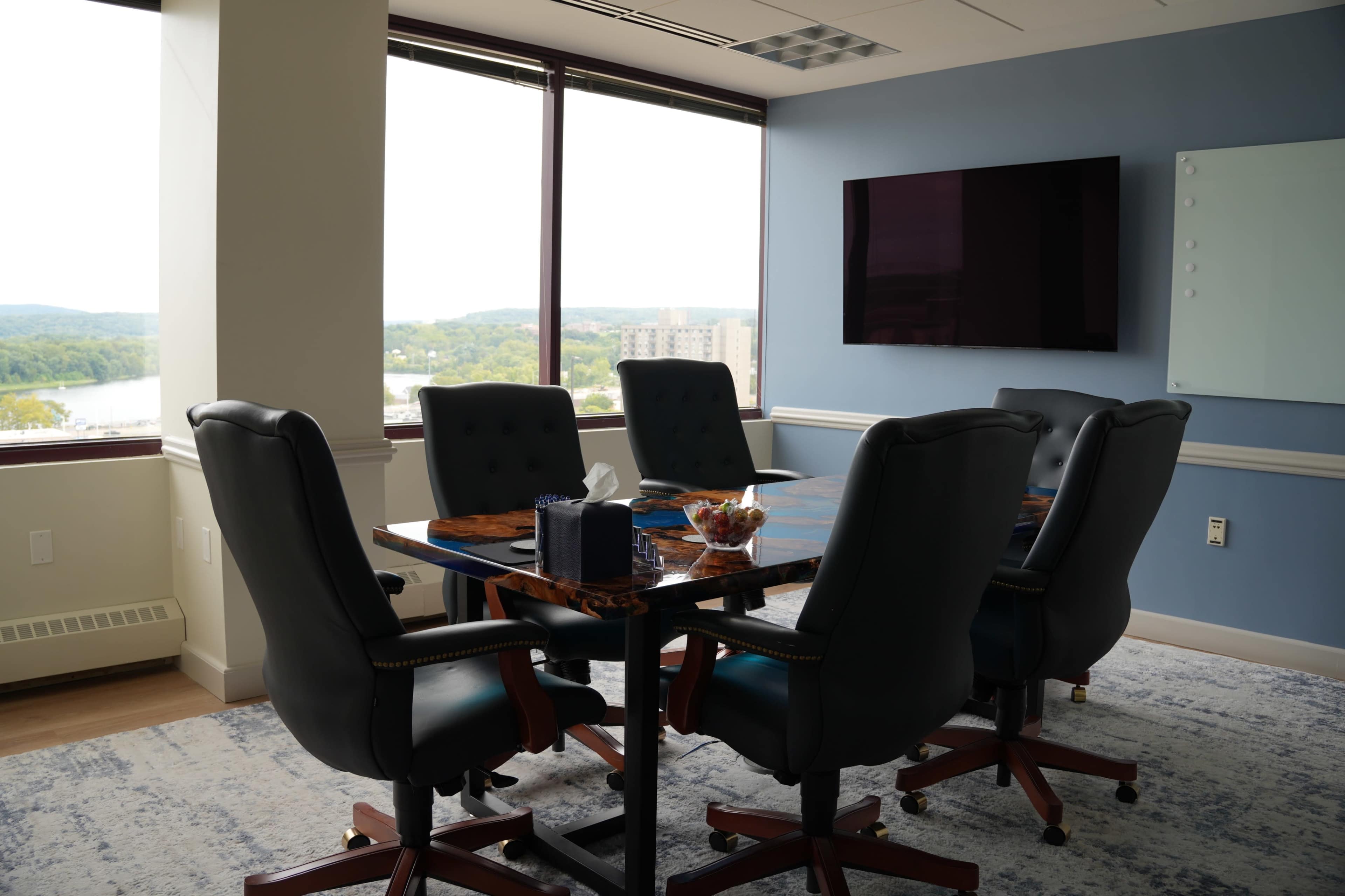The image shows a conference room with a large table surrounded by six black leather chairs, a wall-mounted television, and windows offering a view of the landscape outside.