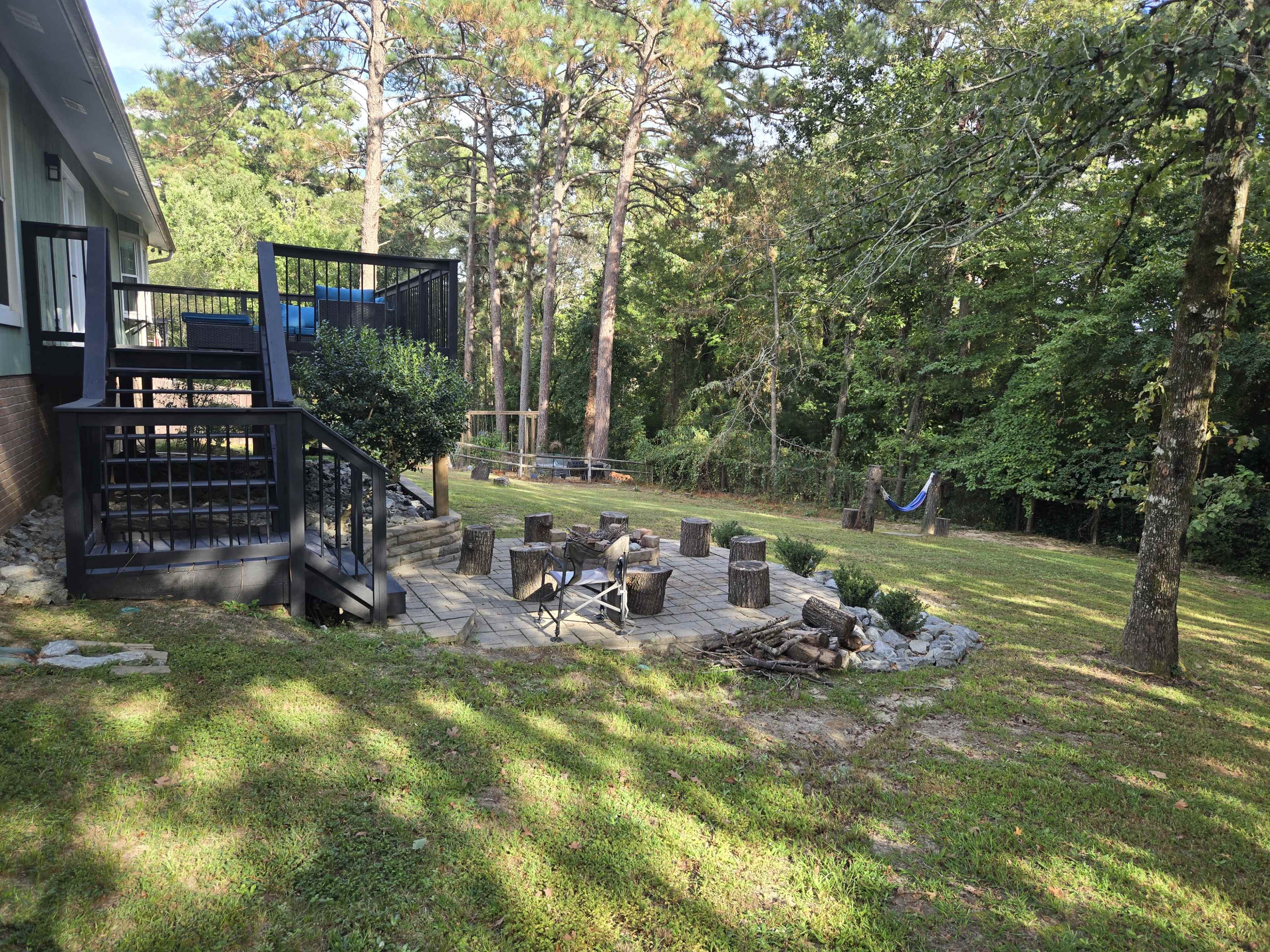 The image shows a backyard area with a stone fire pit surrounded by wooden logs, a hammock hanging between trees, and a deck leading to a house.