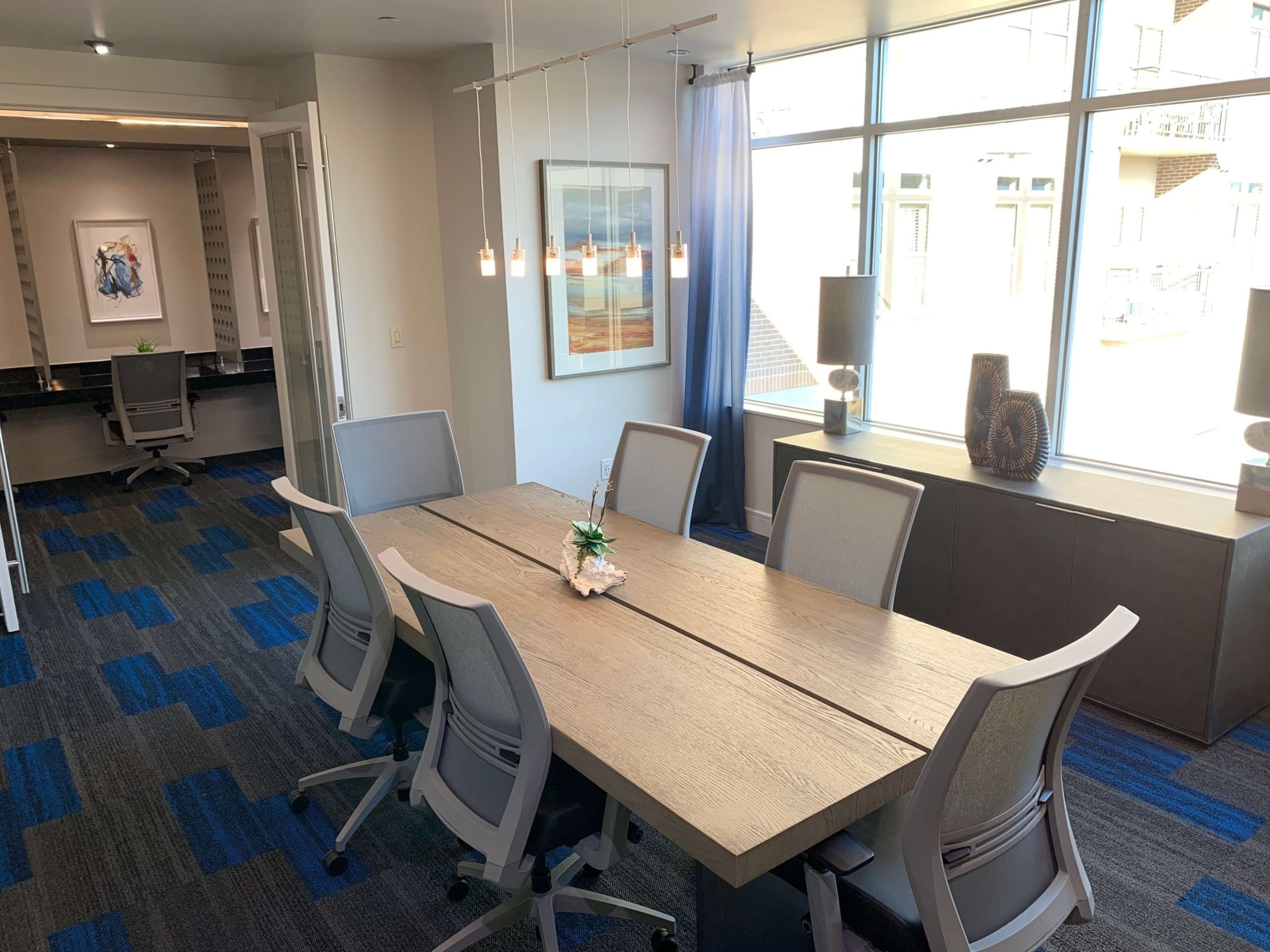 A conference room features a large wooden table surrounded by ergonomic chairs, with large windows providing natural light and a modern decor.