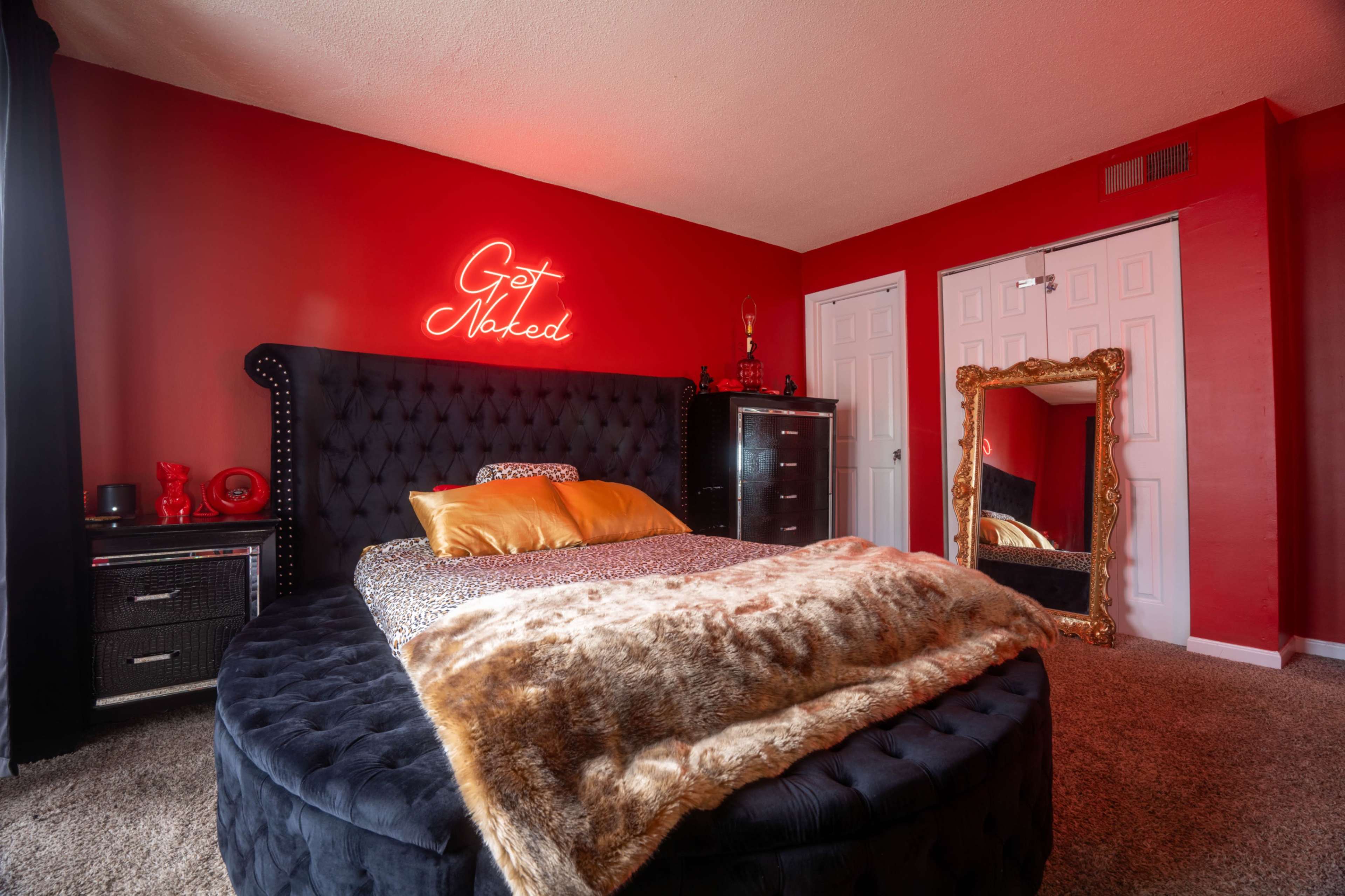 The image shows a bedroom with a round black bed featuring a fur throw, a neon sign that says "Get Naked," and red walls.