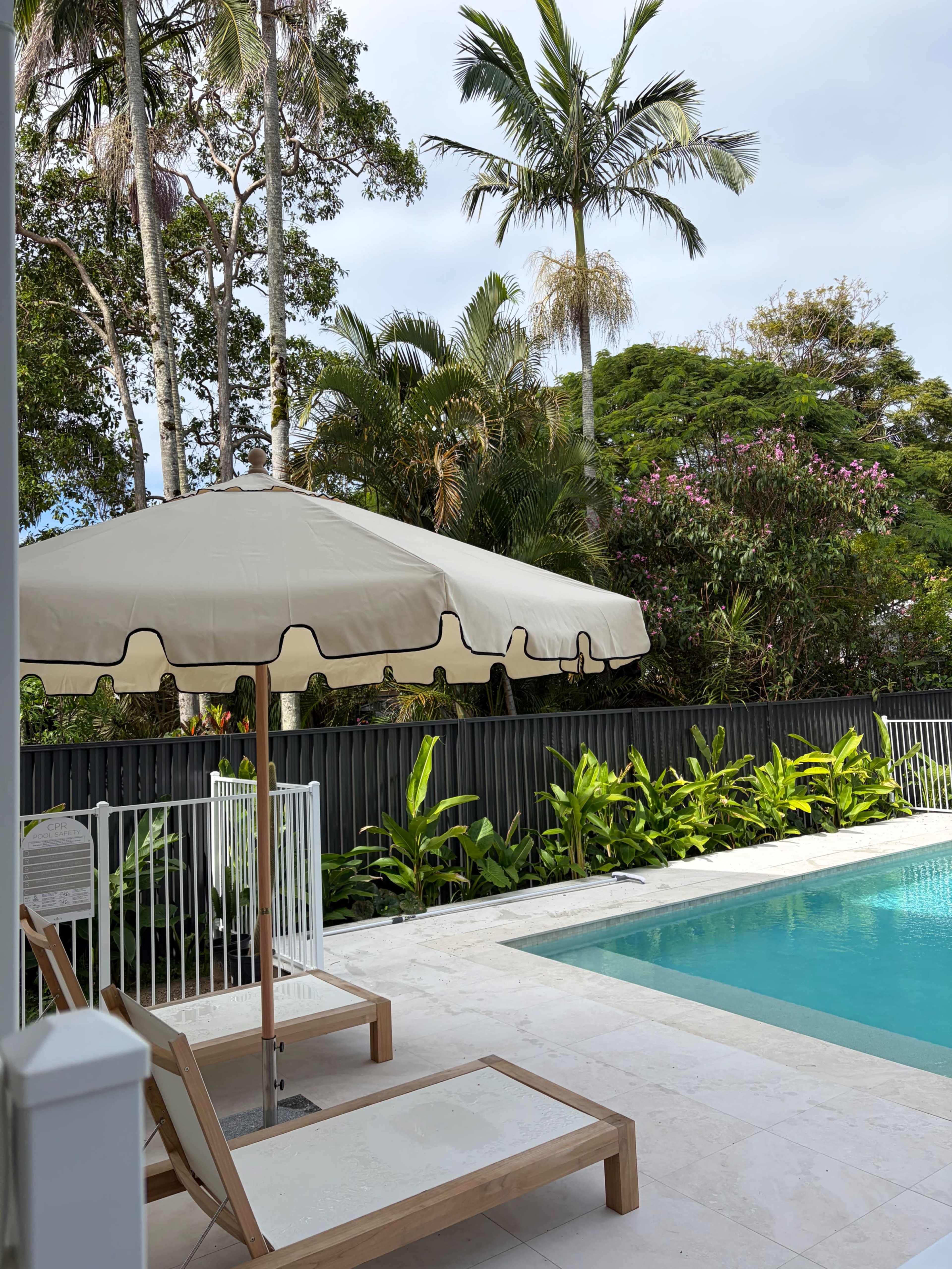 A spacious outdoor area features a swimming pool surrounded by lush greenery and tall palm trees, with a sunshade above a pair of wooden lounge chairs.