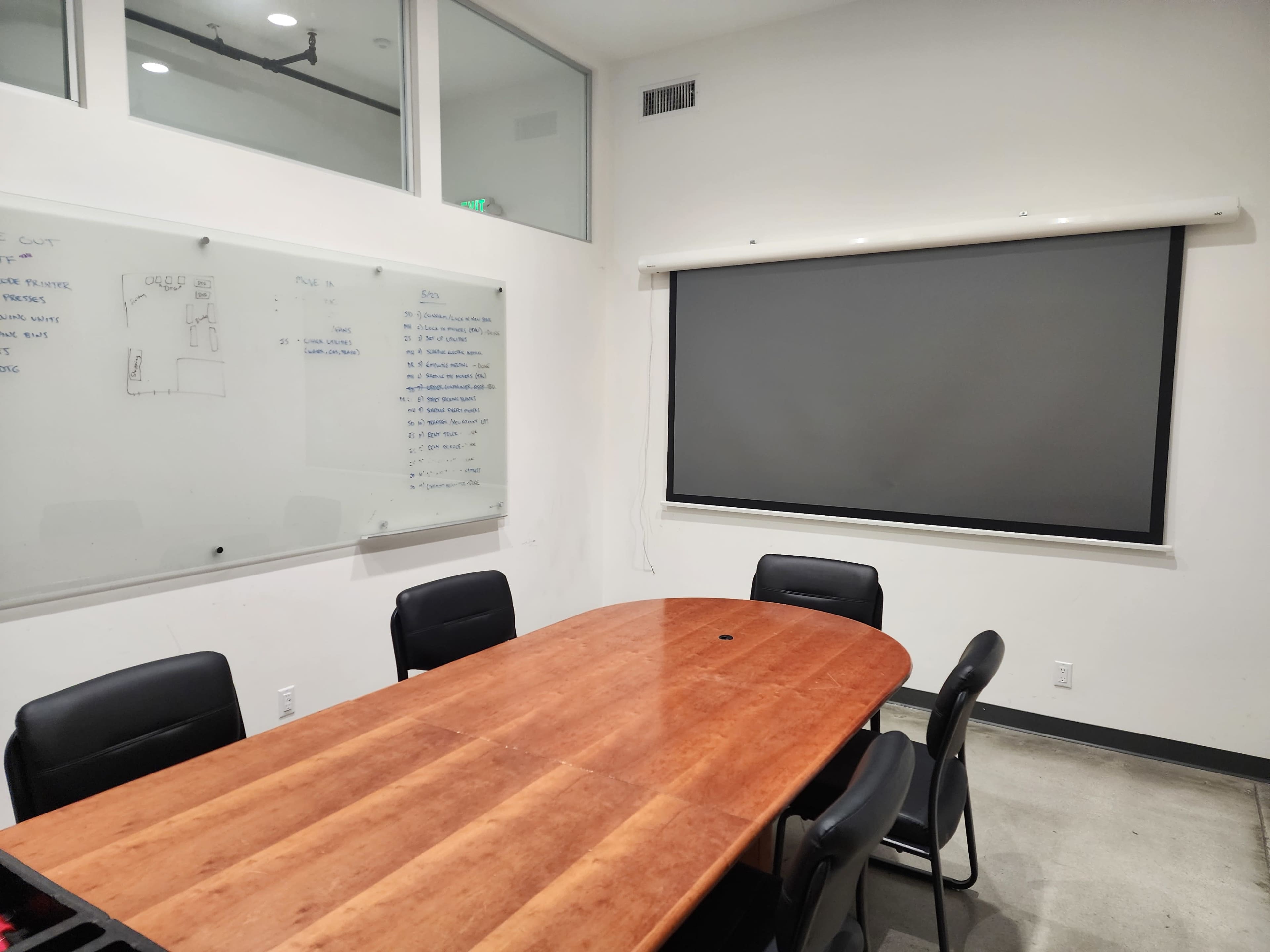 A conference room features a large wooden table surrounded by black chairs, a whiteboard on one wall, and a projection screen on another wall.