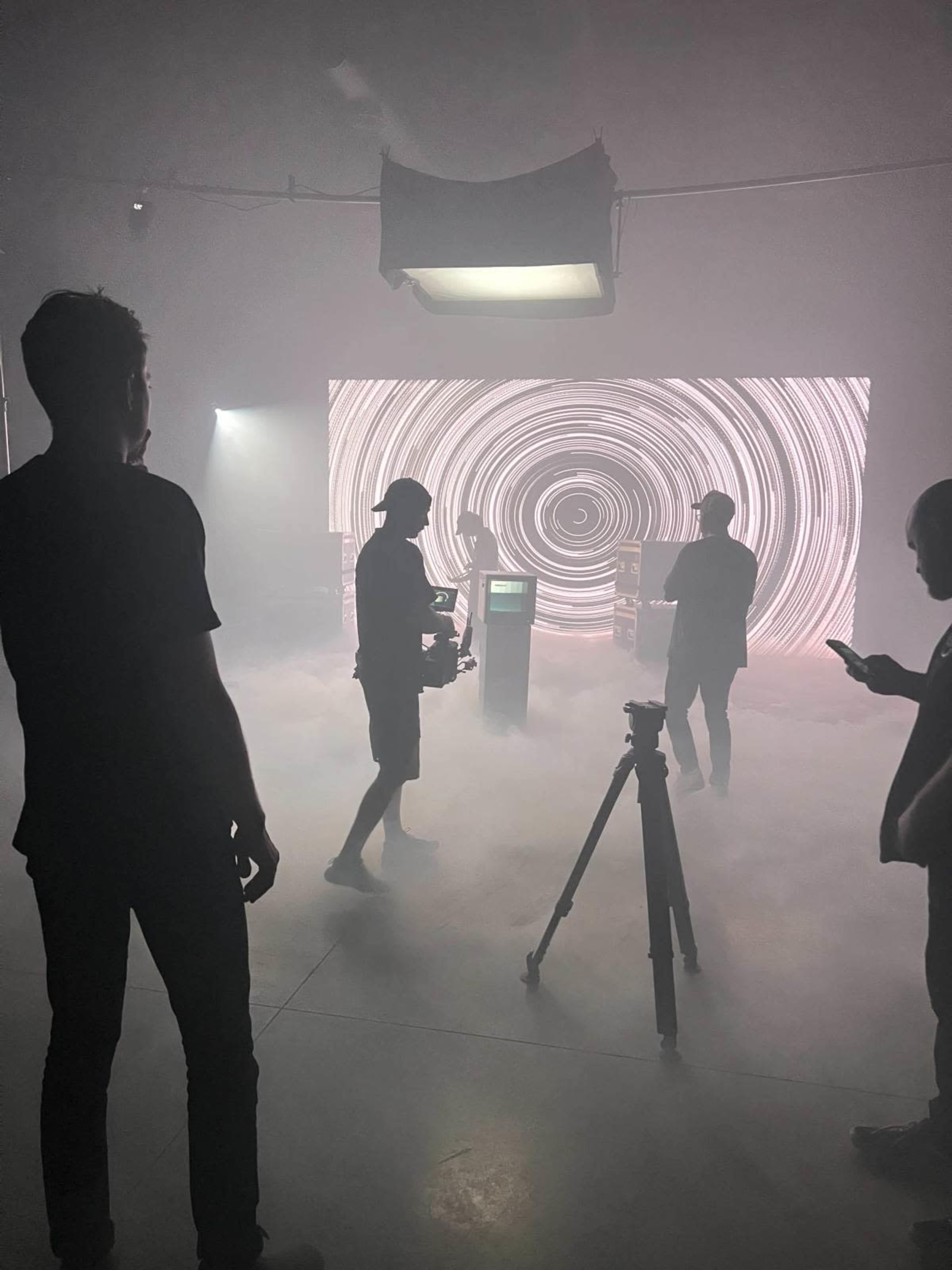 A group of people stands in a dimly lit room with smoke and a large, swirling light display in the background.