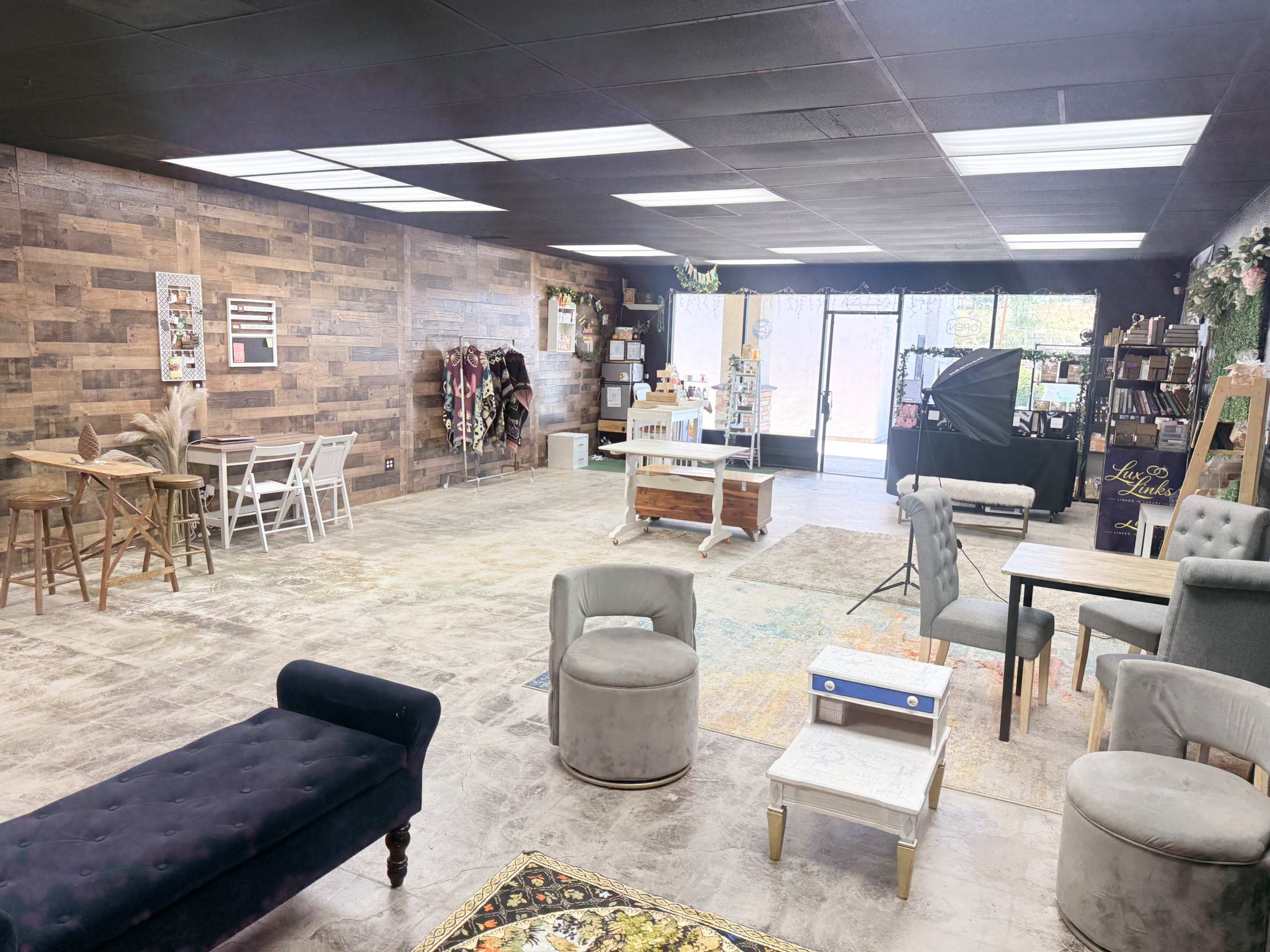 The image shows a spacious, empty room with wooden walls, various types of seating, tables, and decorative elements, including a backdrop setup.