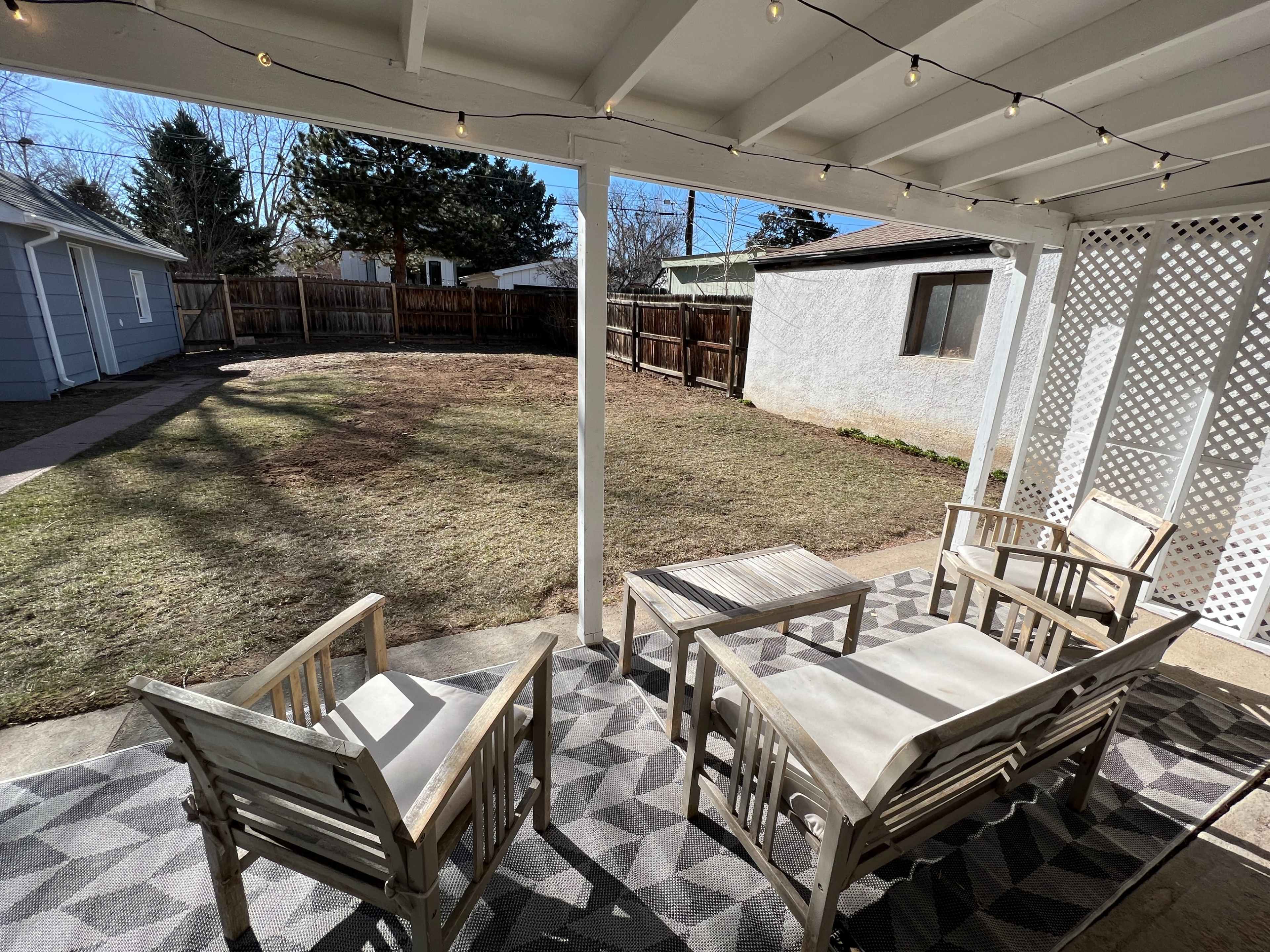 A covered patio with wooden furniture overlooks a grassy yard enclosed by a wooden fence.
