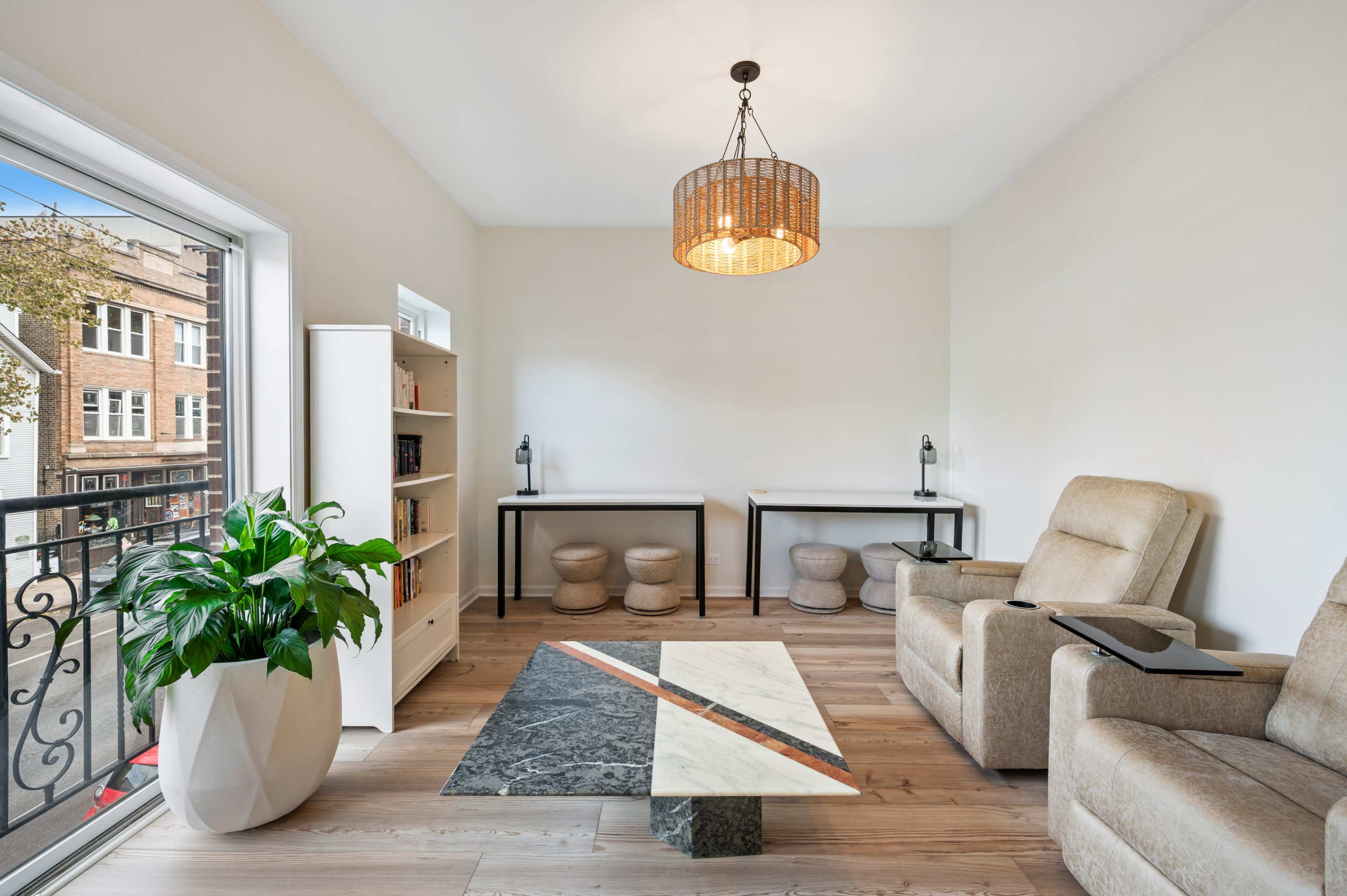 The image shows a minimalist living room featuring two lounge chairs, a coffee table with a geometric design, and a large window with a view of a street.