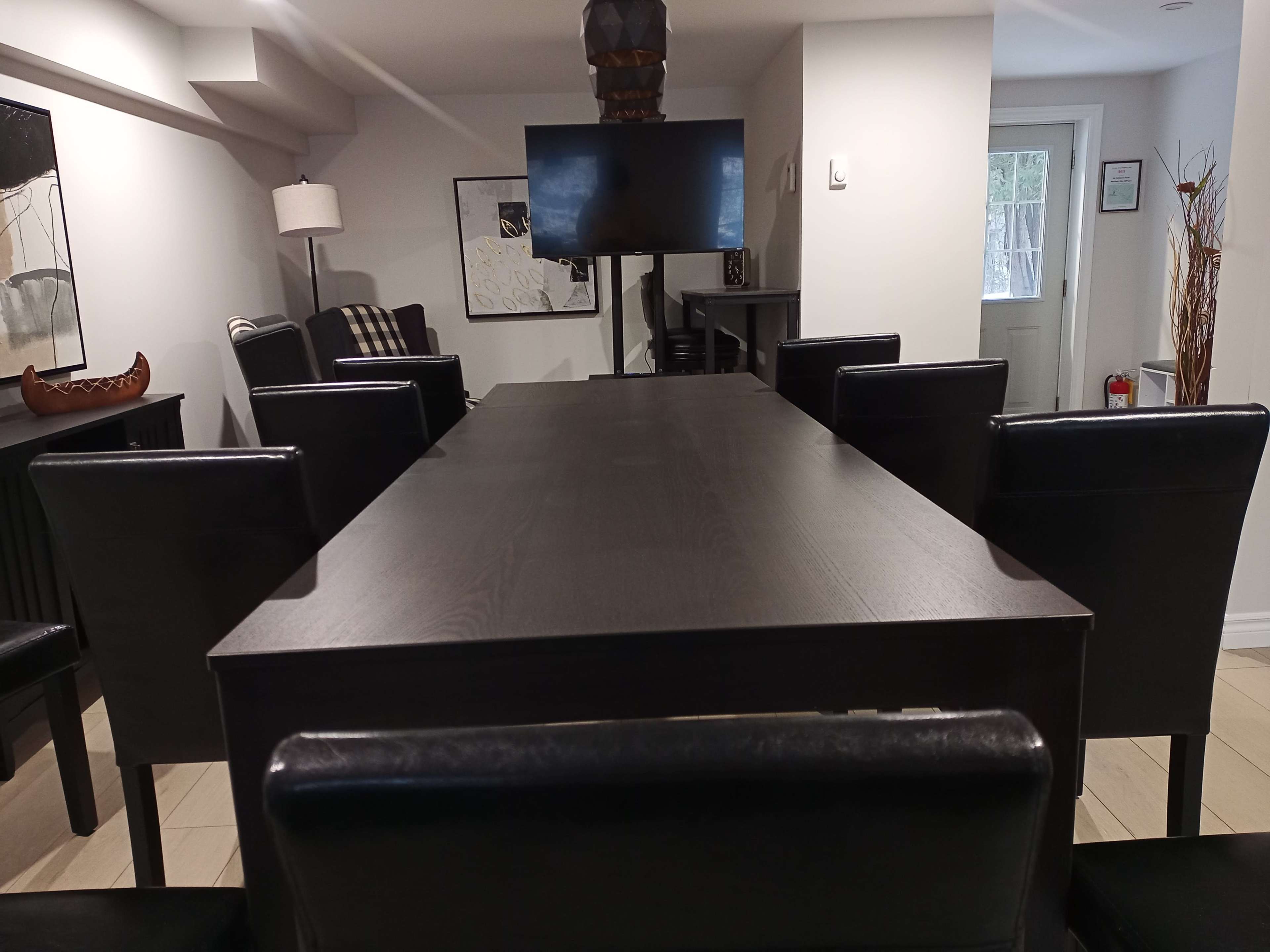 A long wooden dining table with black chairs is positioned in a well-lit room featuring a television and various decorations.