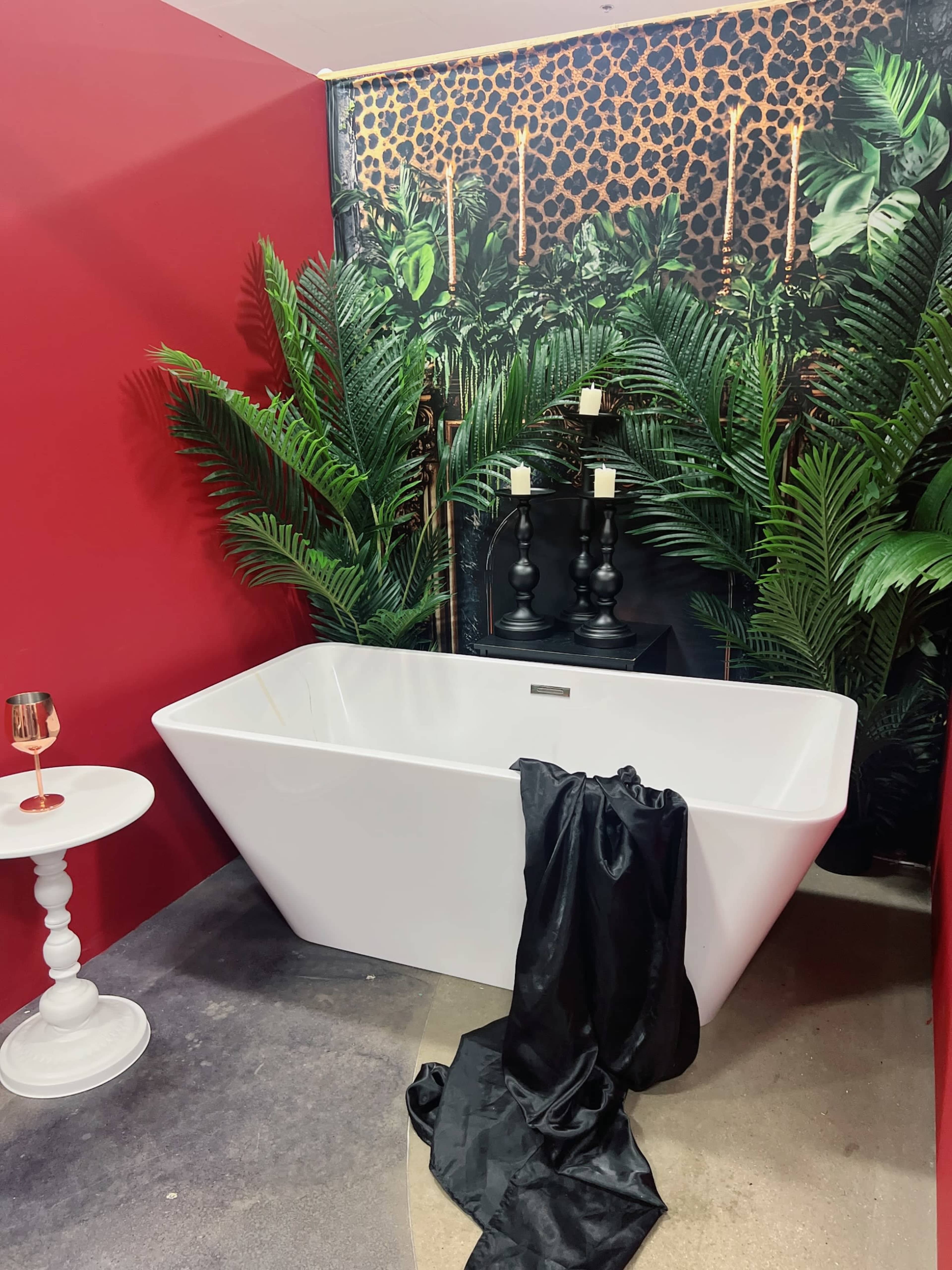 The image features a modern white bathtub with a black drape, surrounded by lush green plants against a red wall and a decorative backdrop.