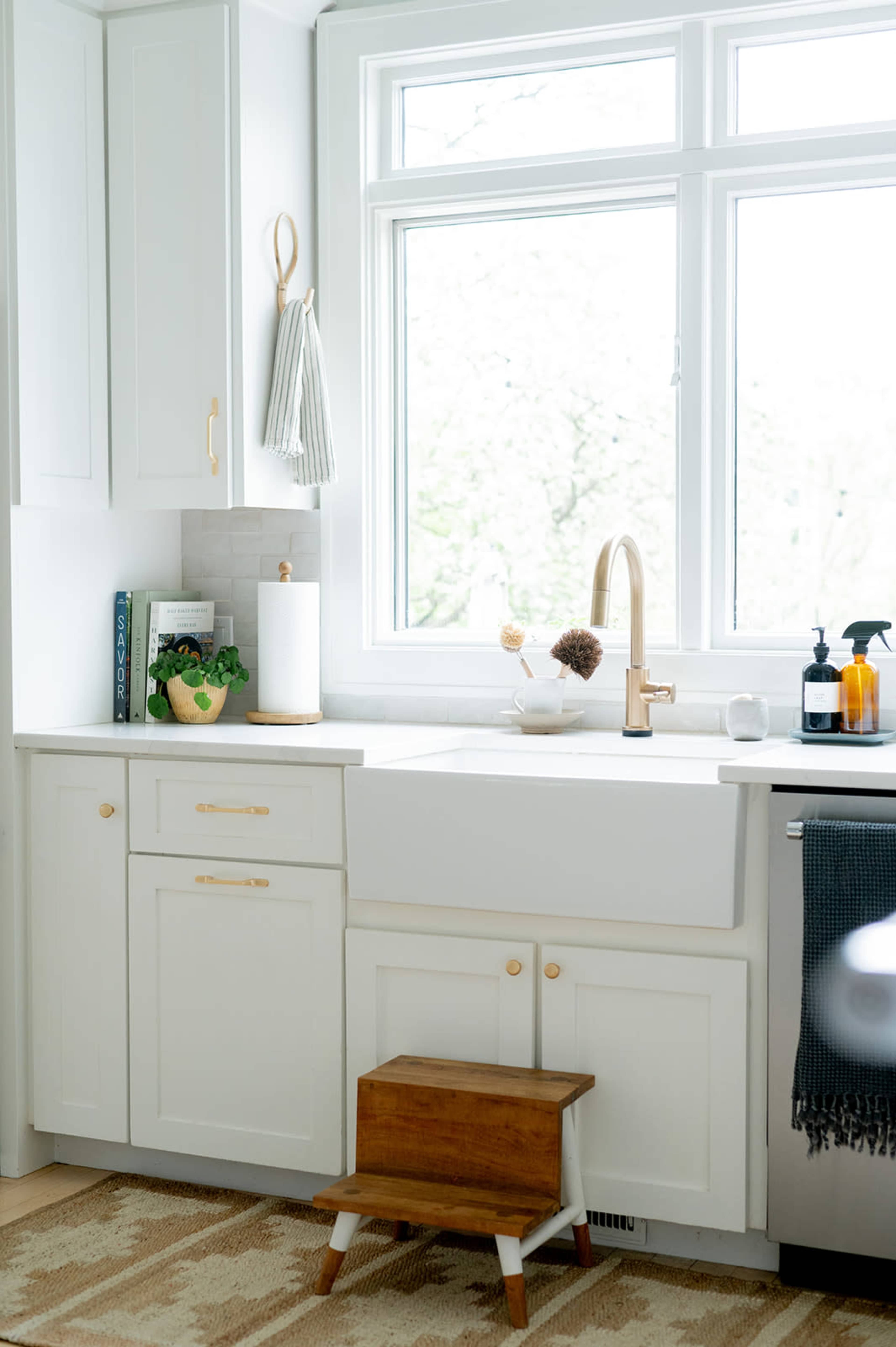 A bright kitchen features white cabinetry, a farmhouse sink, a wooden step stool, and a window with a view of greenery outside.