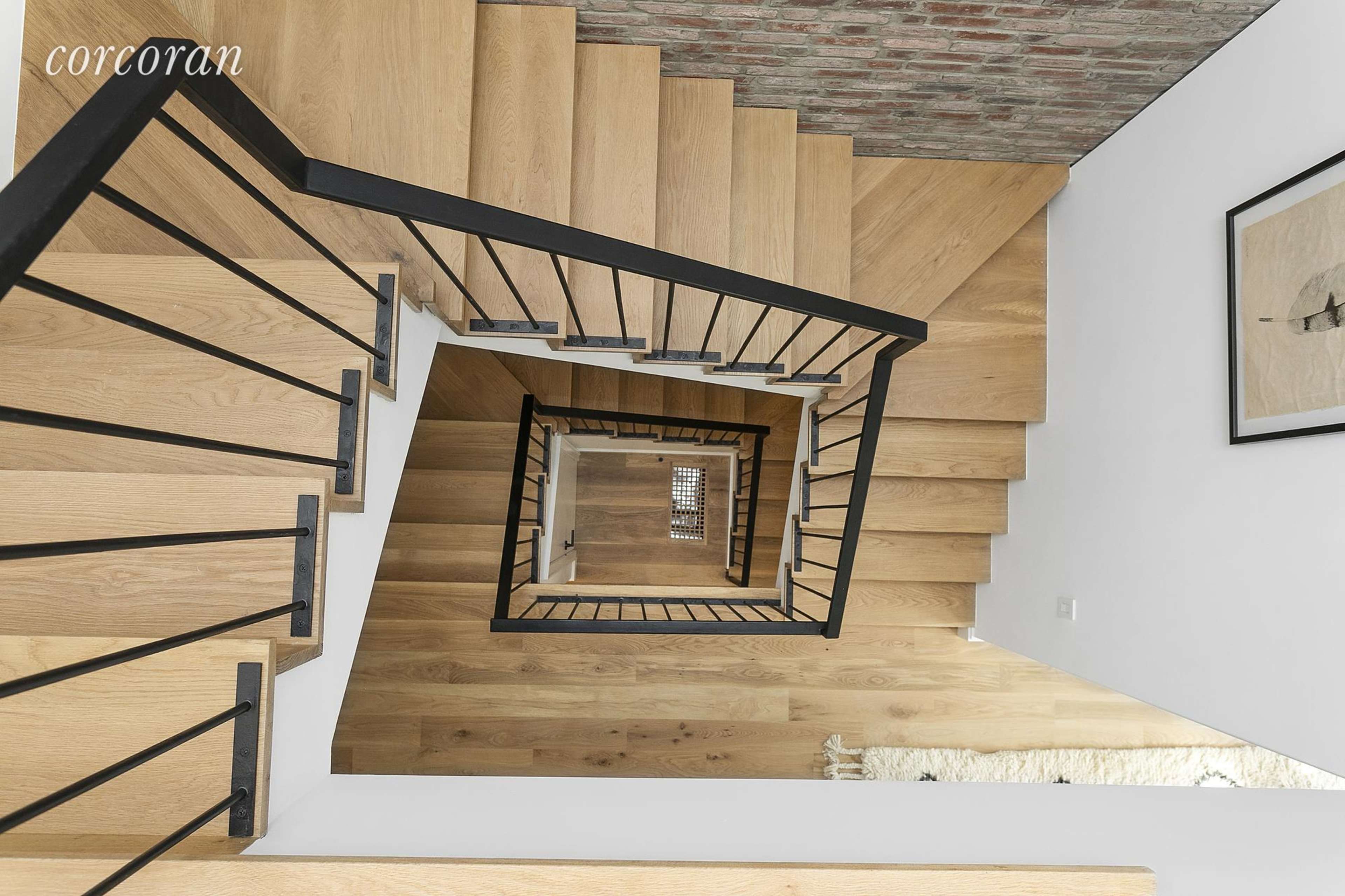 A downward view of a wooden staircase with black railings, surrounded by a brick wall and light-colored flooring.