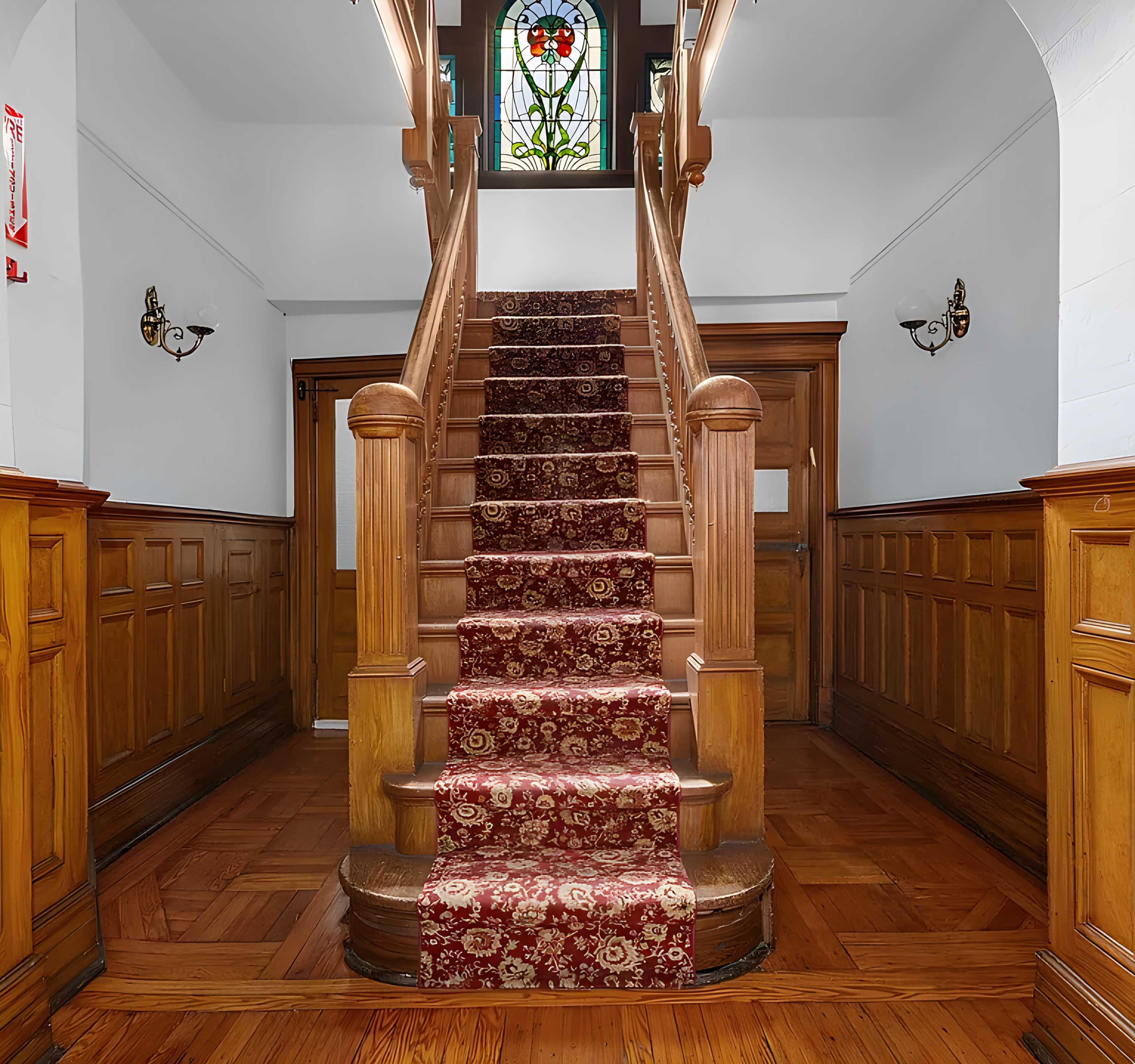A wooden staircase with a patterned carpet runner, leading up to a stained glass window in a well-lit entryway.