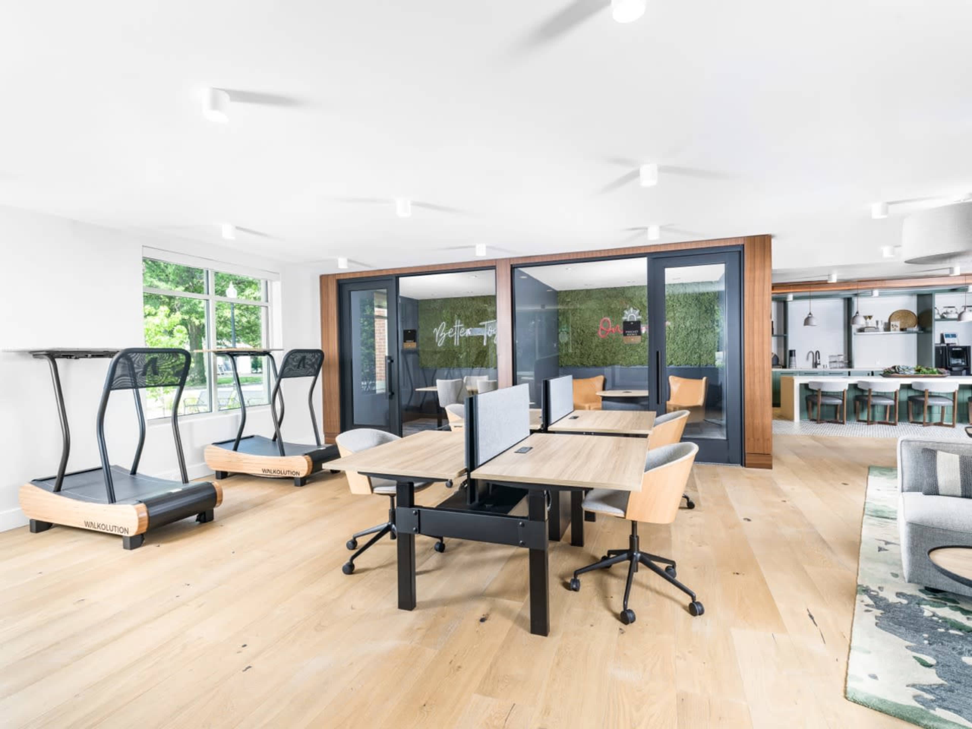 The image shows a modern office space featuring standing desks, a treadmill workstation, and a communal area with a dining table.