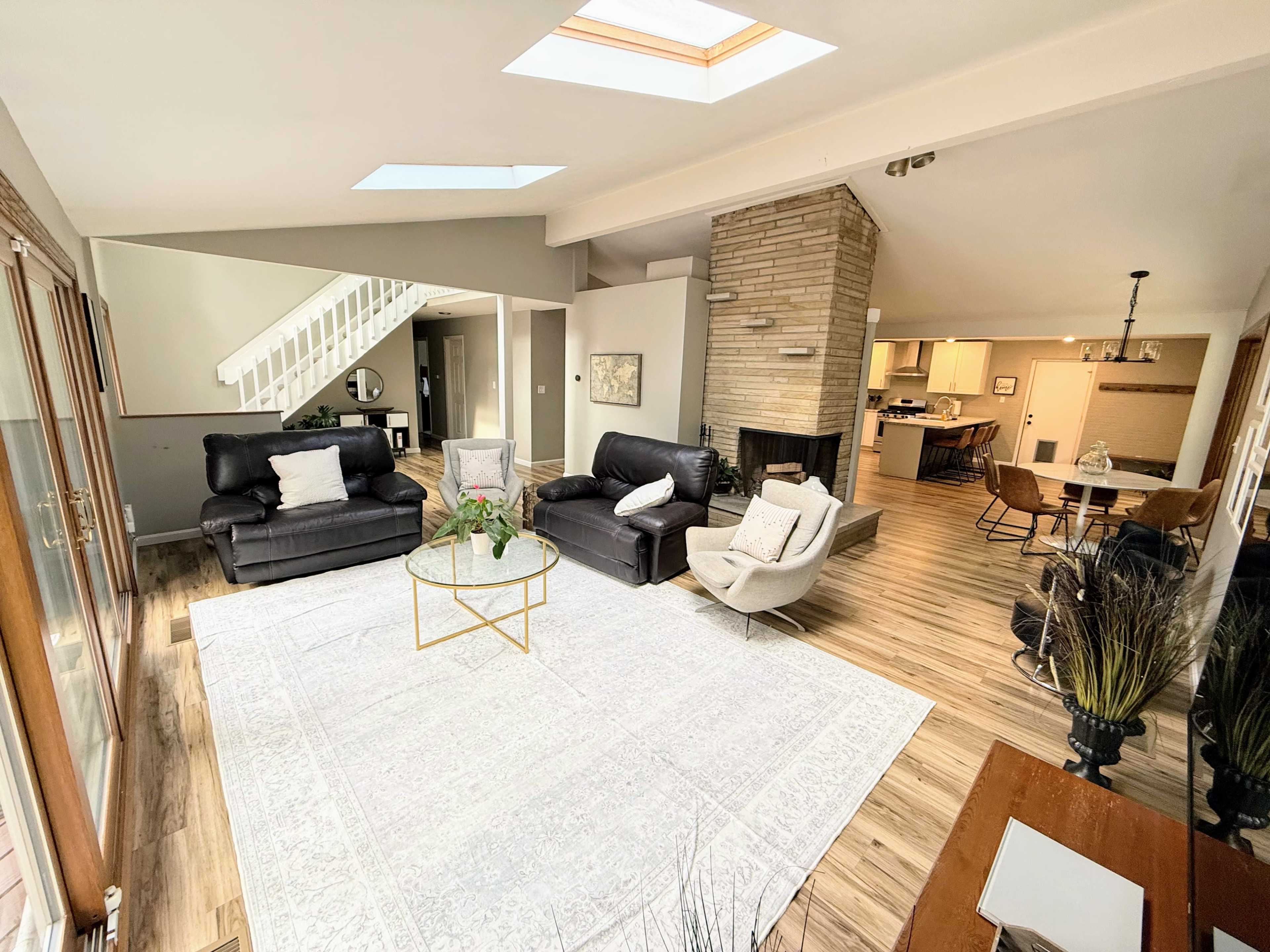 The living room features two black leather sofas and a glass coffee table centered on a large area rug, with a skylight above and an open layout leading to a dining area in the background.