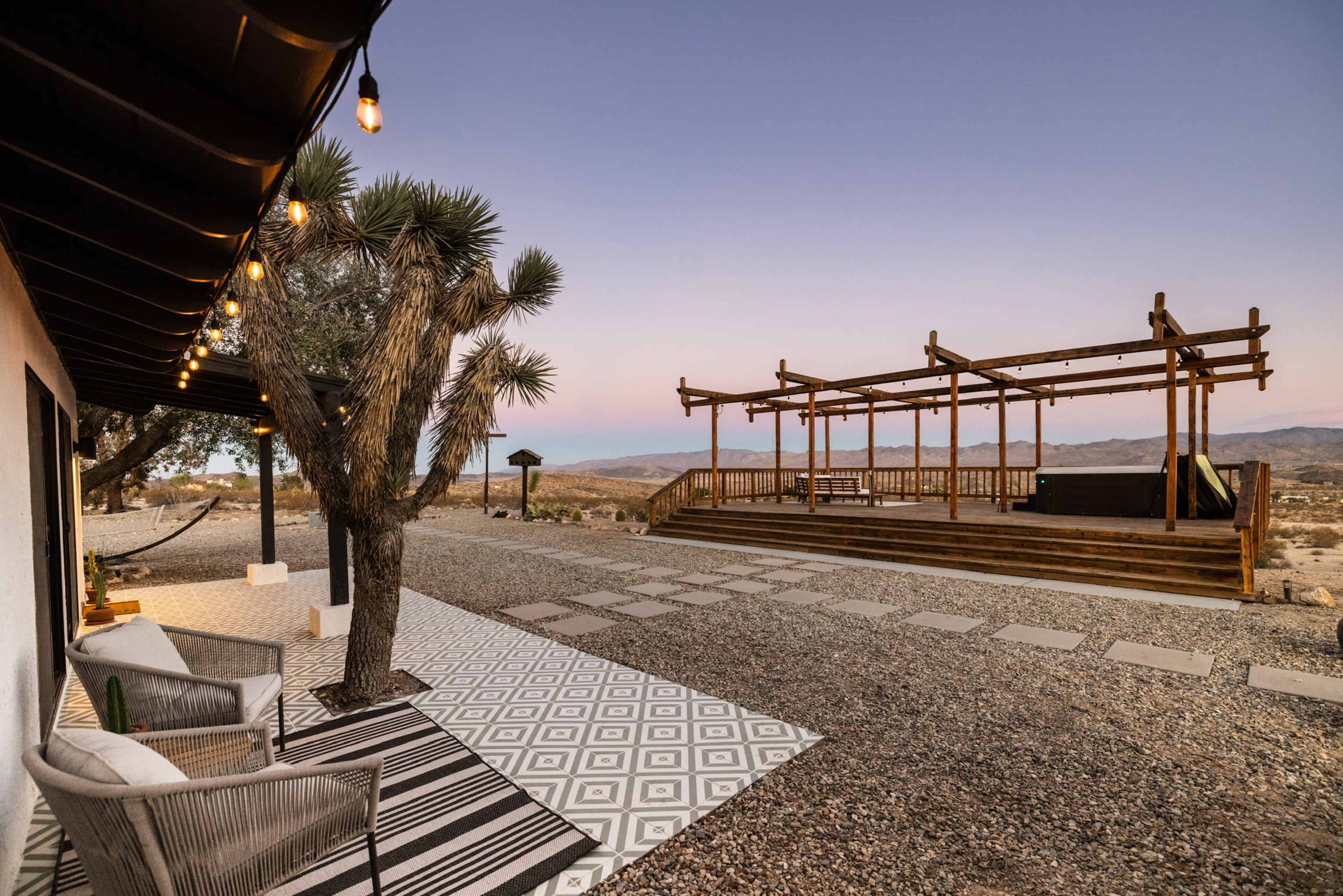 A wooden stage is positioned near a gravel area with patterned pavers and outdoor seating under string lights, surrounded by desert scenery.