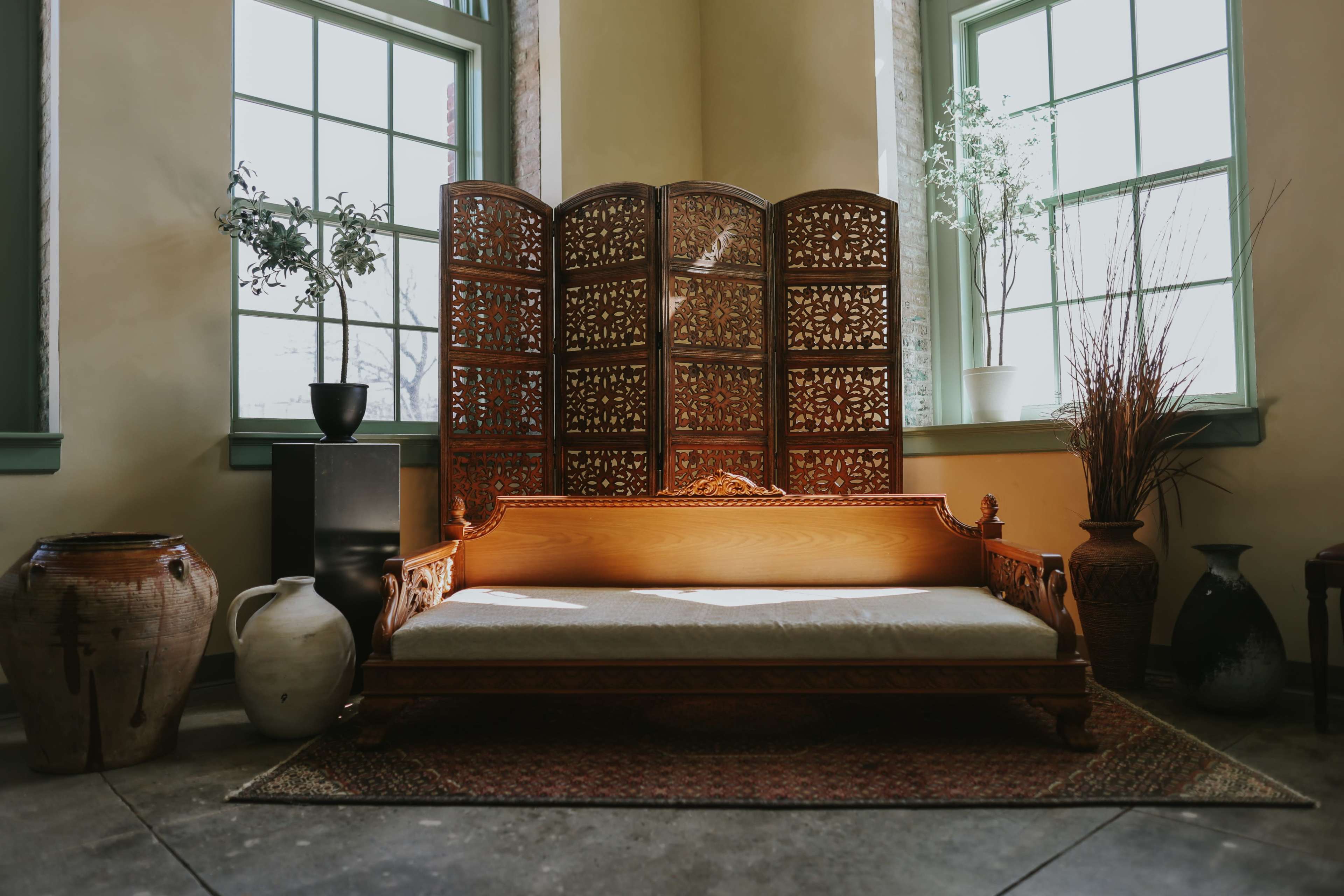 The image features a wooden sofa with intricate carvings set against a backdrop of large windows and a decorative room divider, surrounded by potted plants and various decorative items.