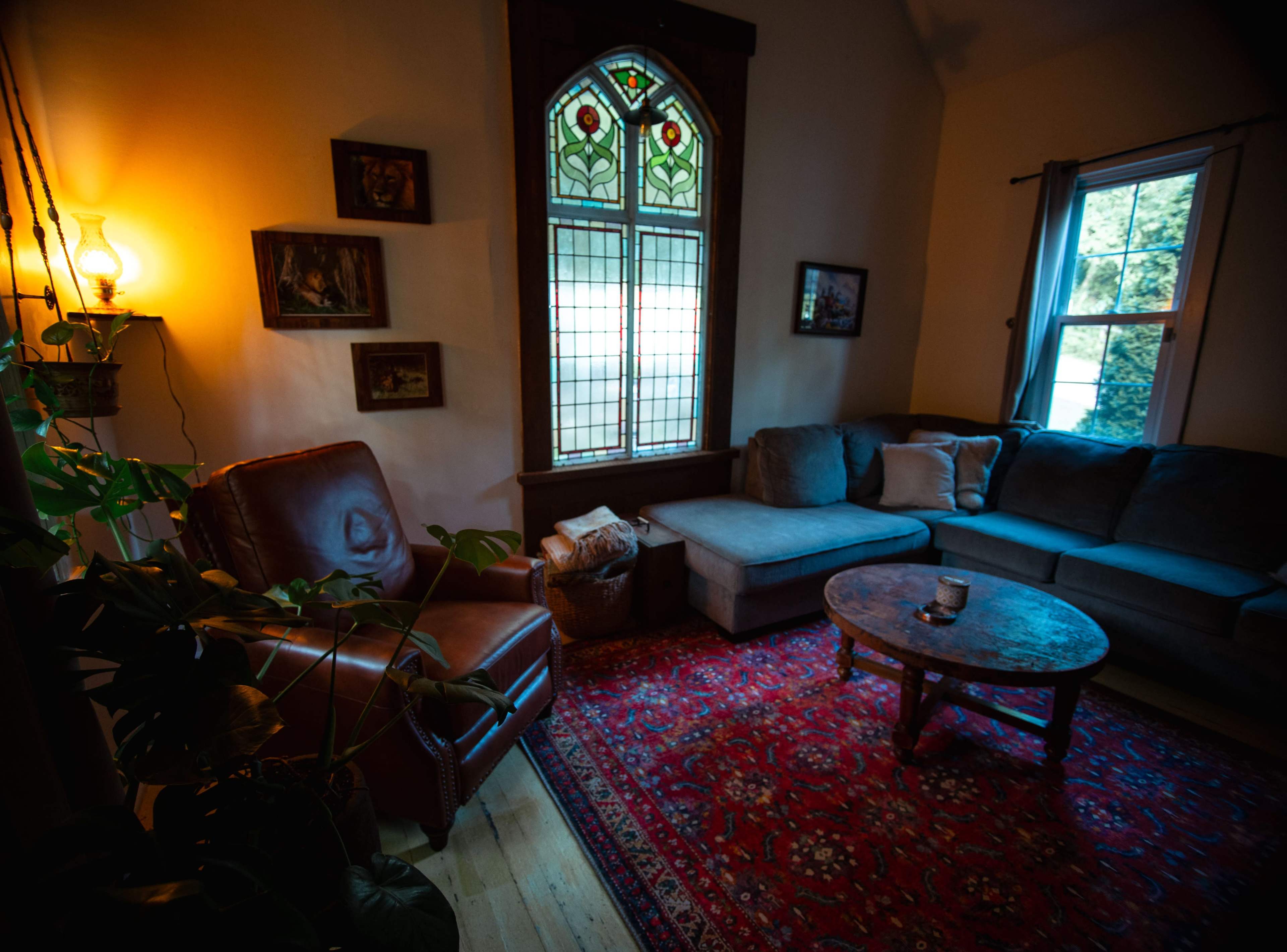 A cozy living room featuring a large sectional sofa, a wooden coffee table, a stained glass window, and a warm light illuminating the space.