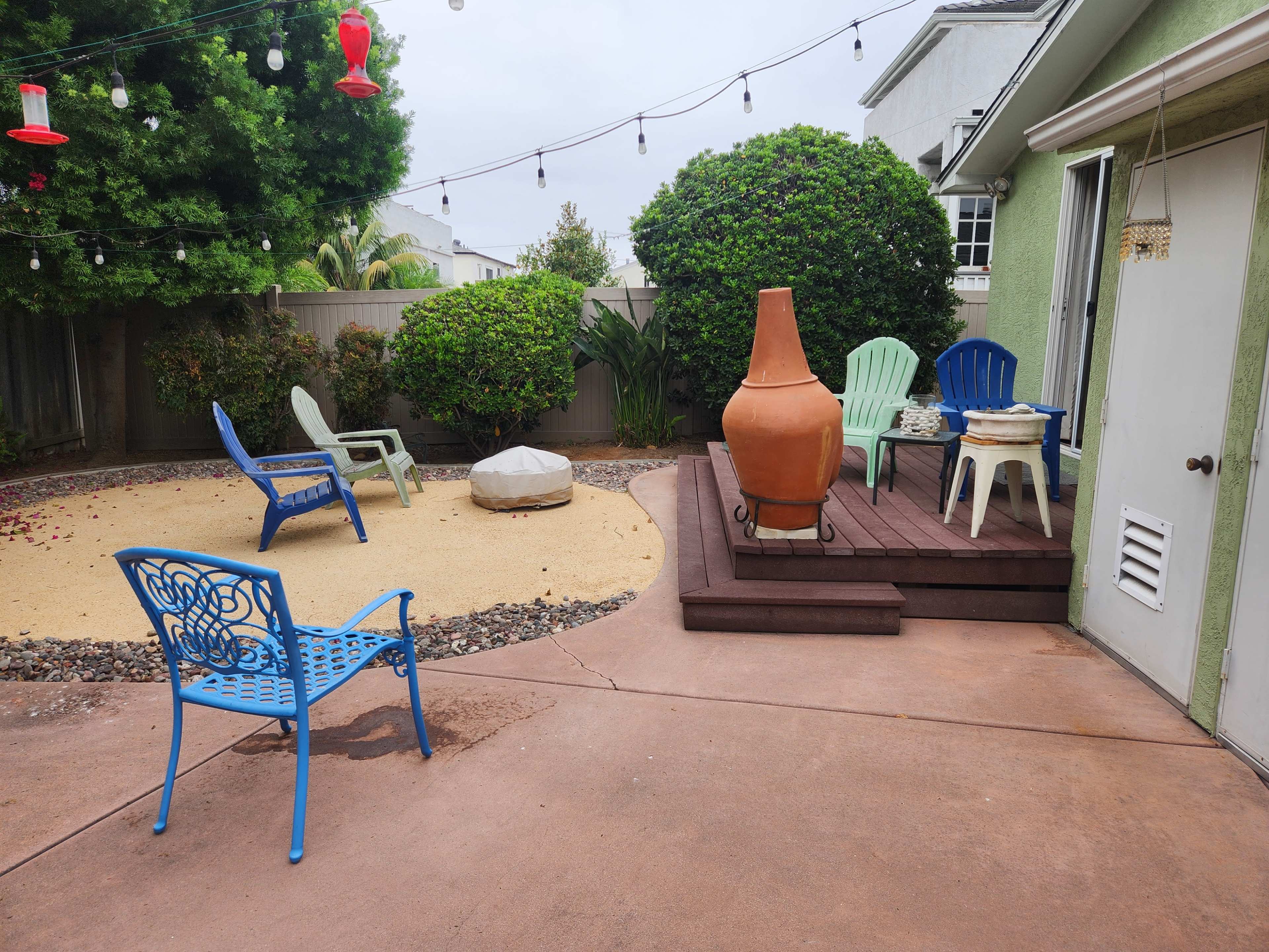 The backyard features a sand area, several chairs, a large terracotta pot, and a wooden deck beside a house.