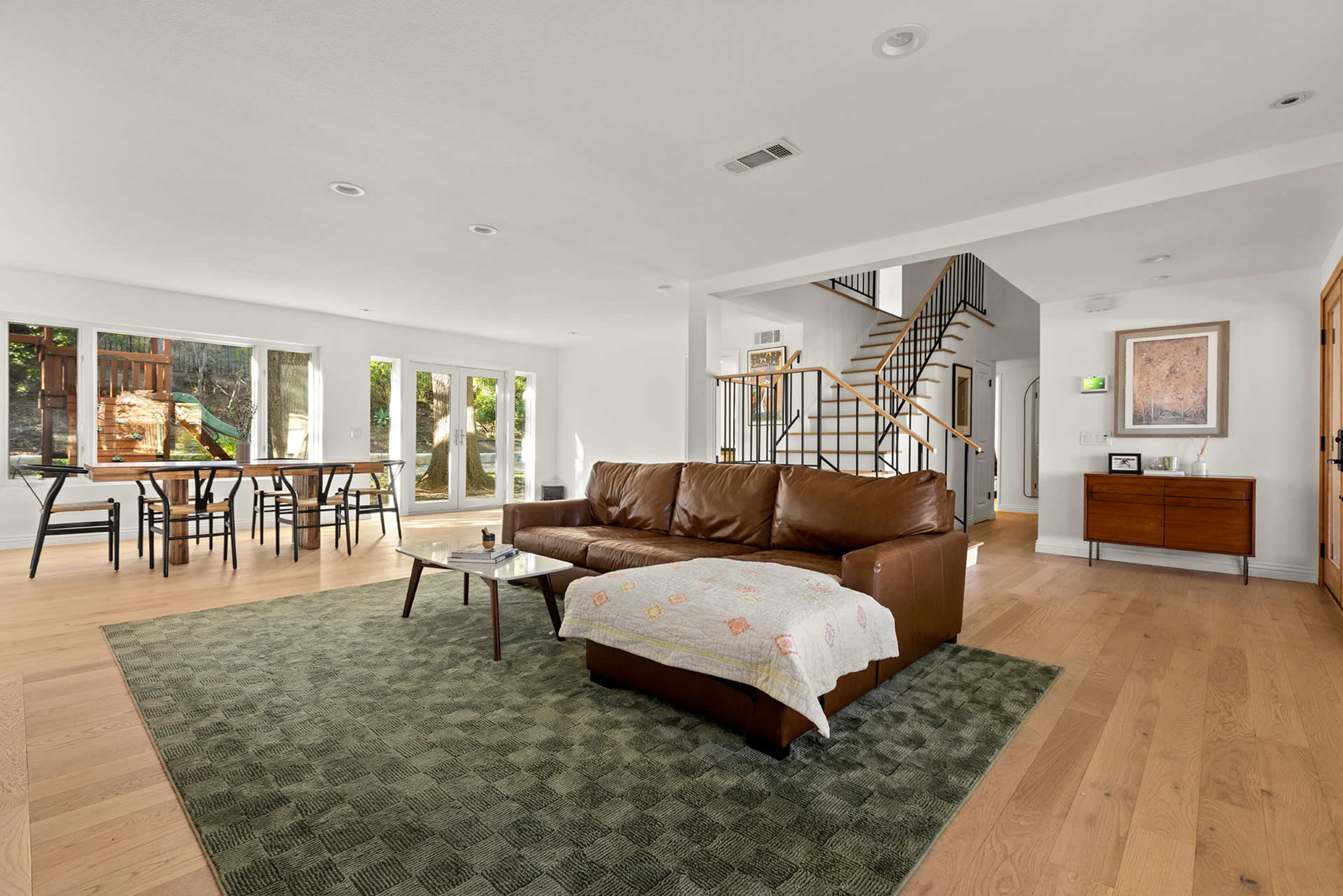 A spacious living room with a brown sofa, a coffee table, and a large area rug, connected to a dining area and a staircase in the background.