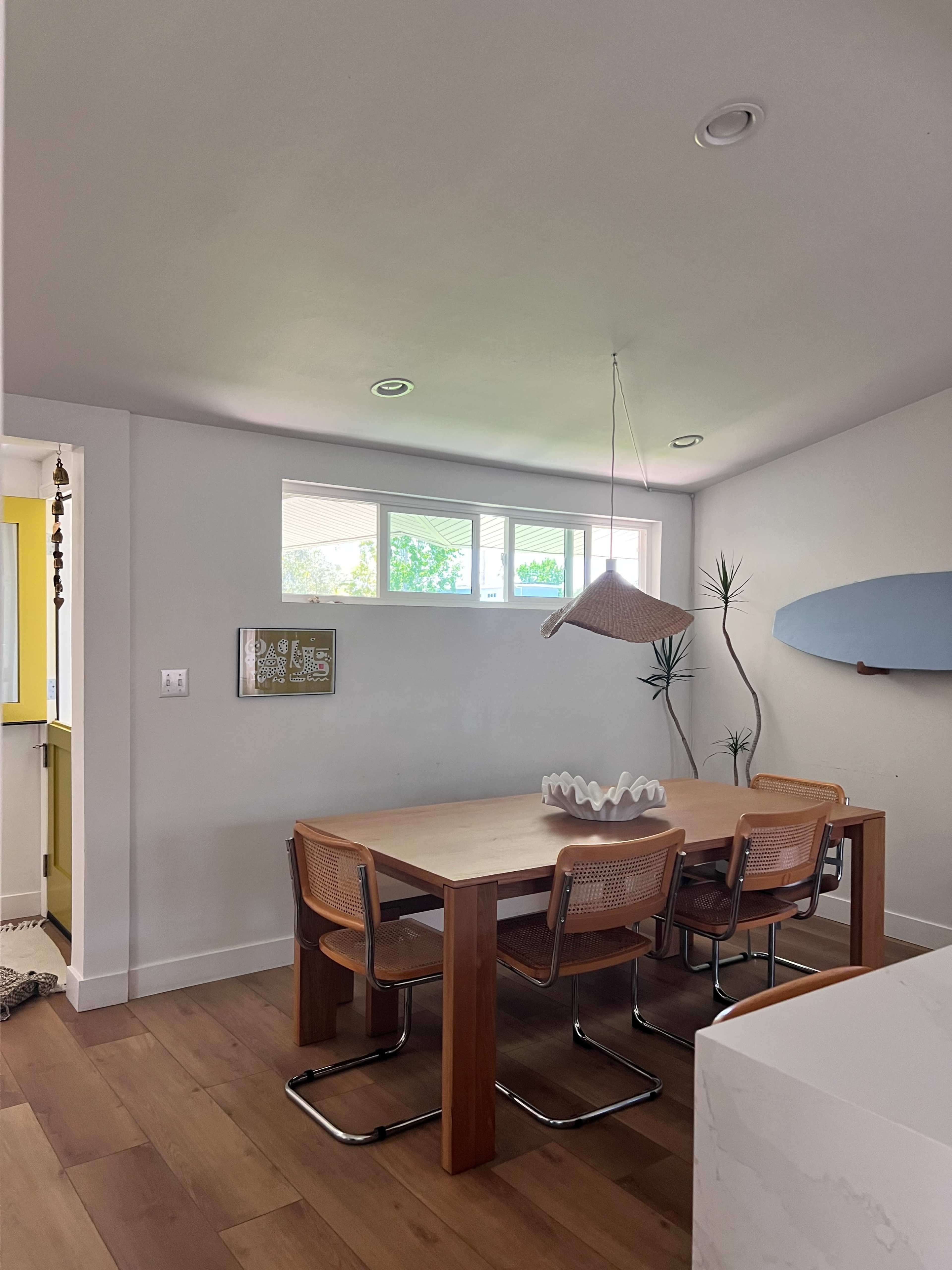 A wooden dining table with metal chairs is positioned beneath a pendant light in a brightly lit room featuring large windows and a potted plant.