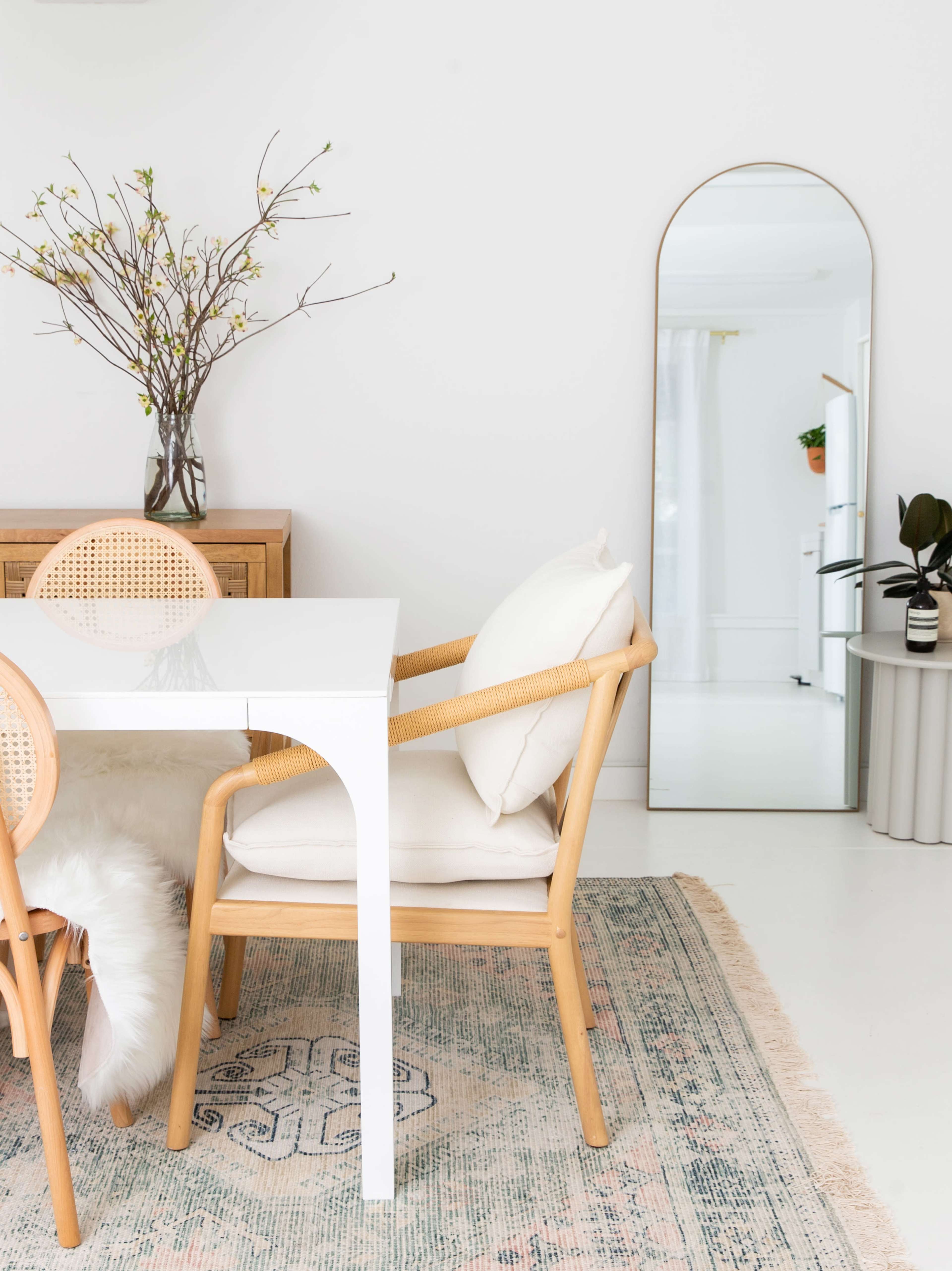 A dining area features a white table with rattan chairs, a flower arrangement in a vase, a large mirror, and a patterned rug.