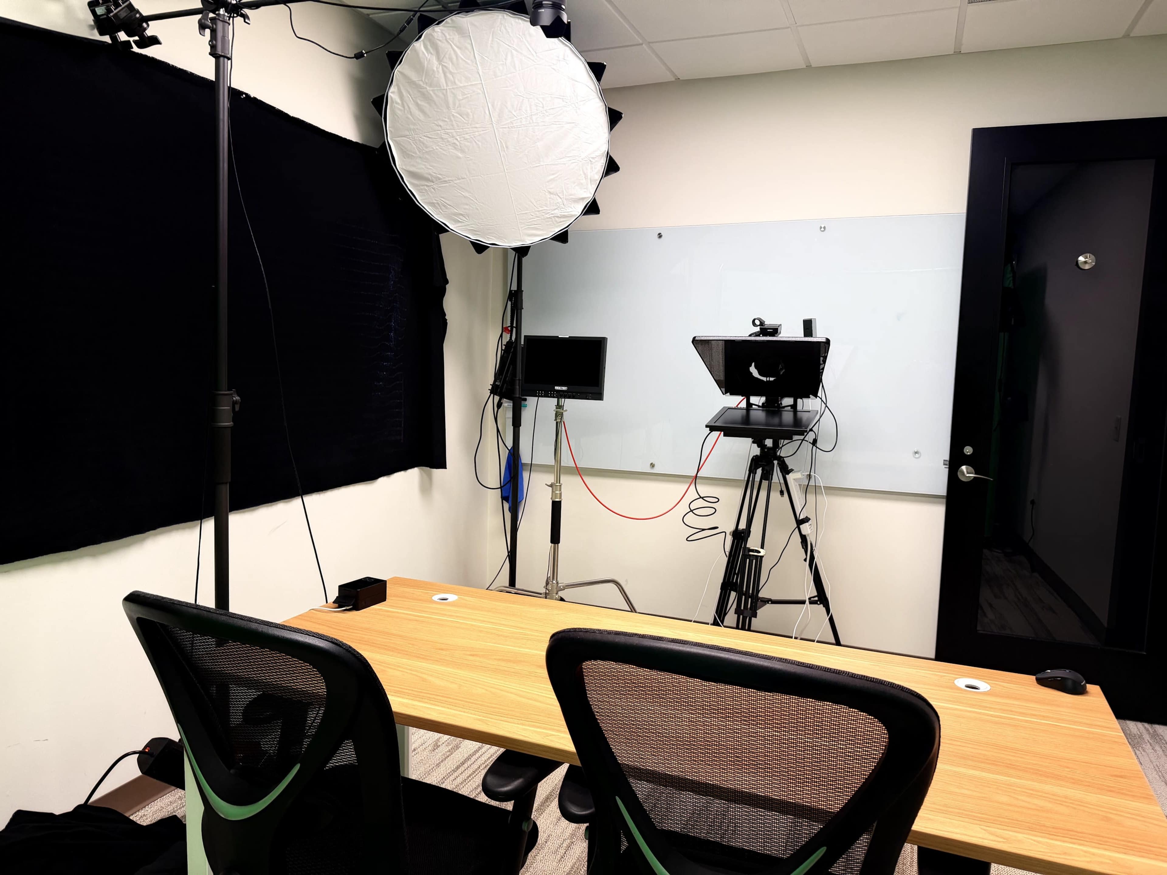 The image shows a small recording studio setup with a wooden table, two chairs, camera equipment, and lighting fixtures arranged against the wall.