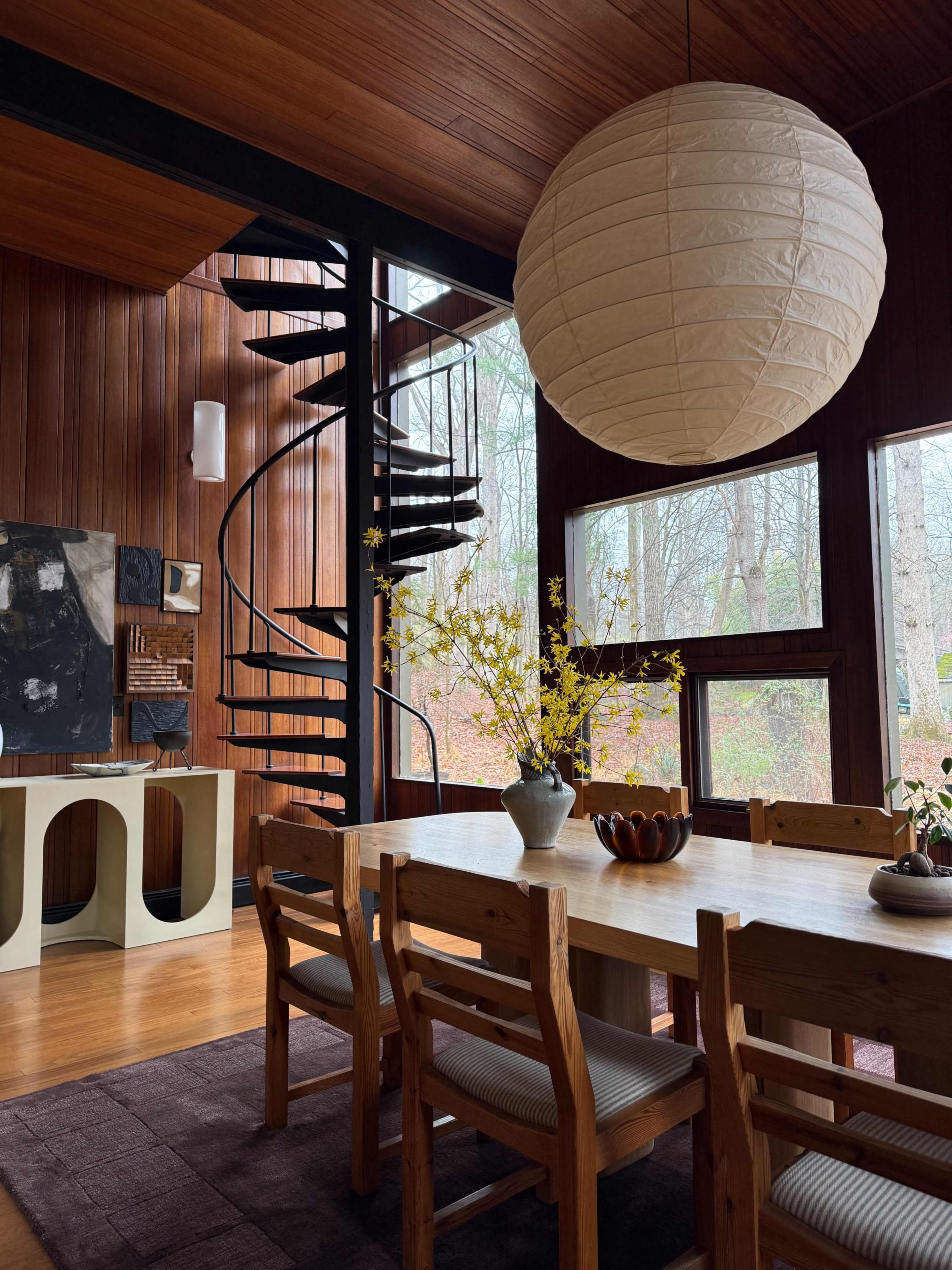 The image depicts a modern interior space featuring a wooden dining area with a table and chairs, a spiral staircase, large windows with views of trees outside, and decorative elements like a vase of yellow flowers.