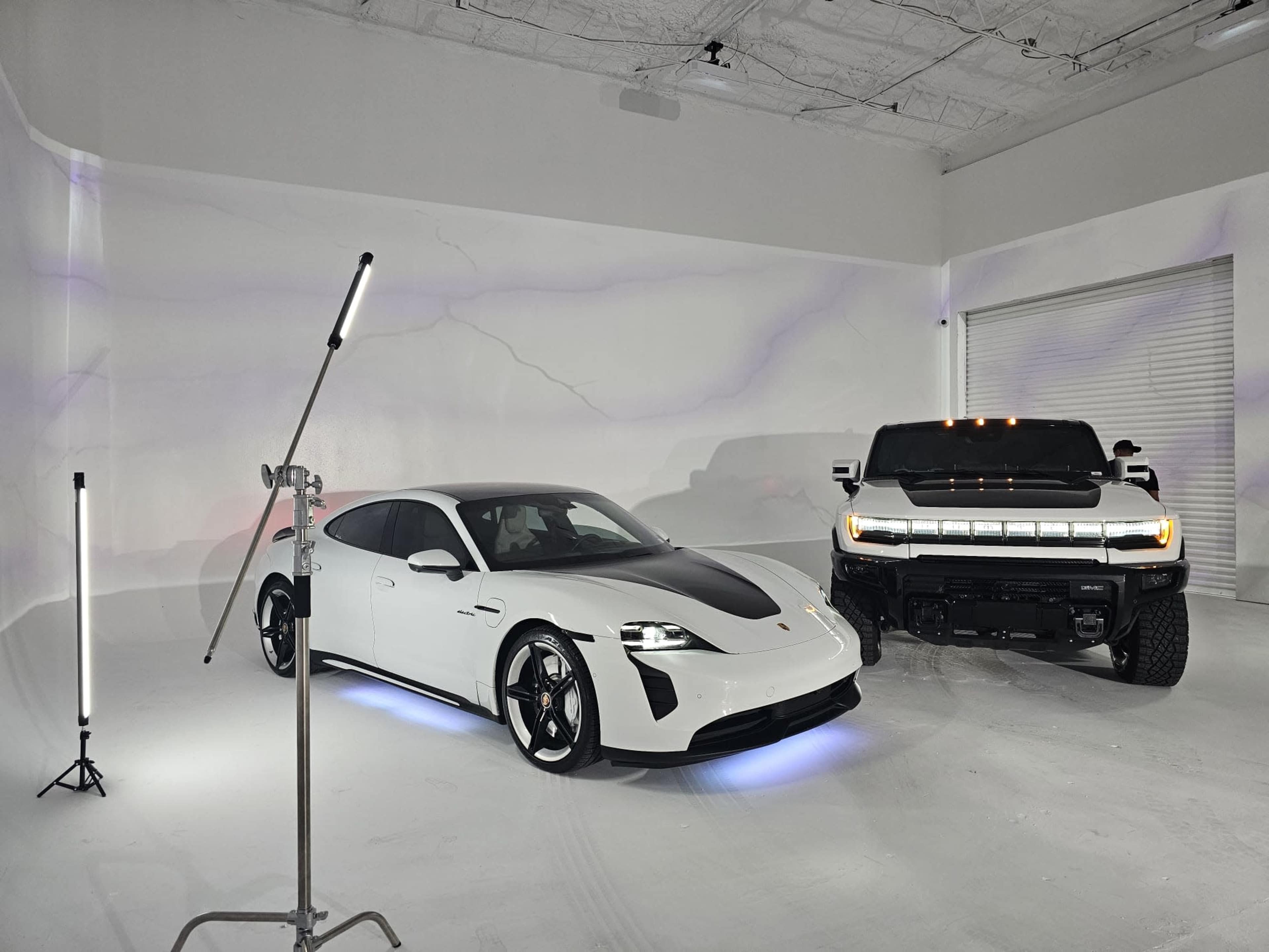 A white Porsche Taycan with a black hood is parked beside a black Hummer EV in a minimalist studio setting with soft lighting.