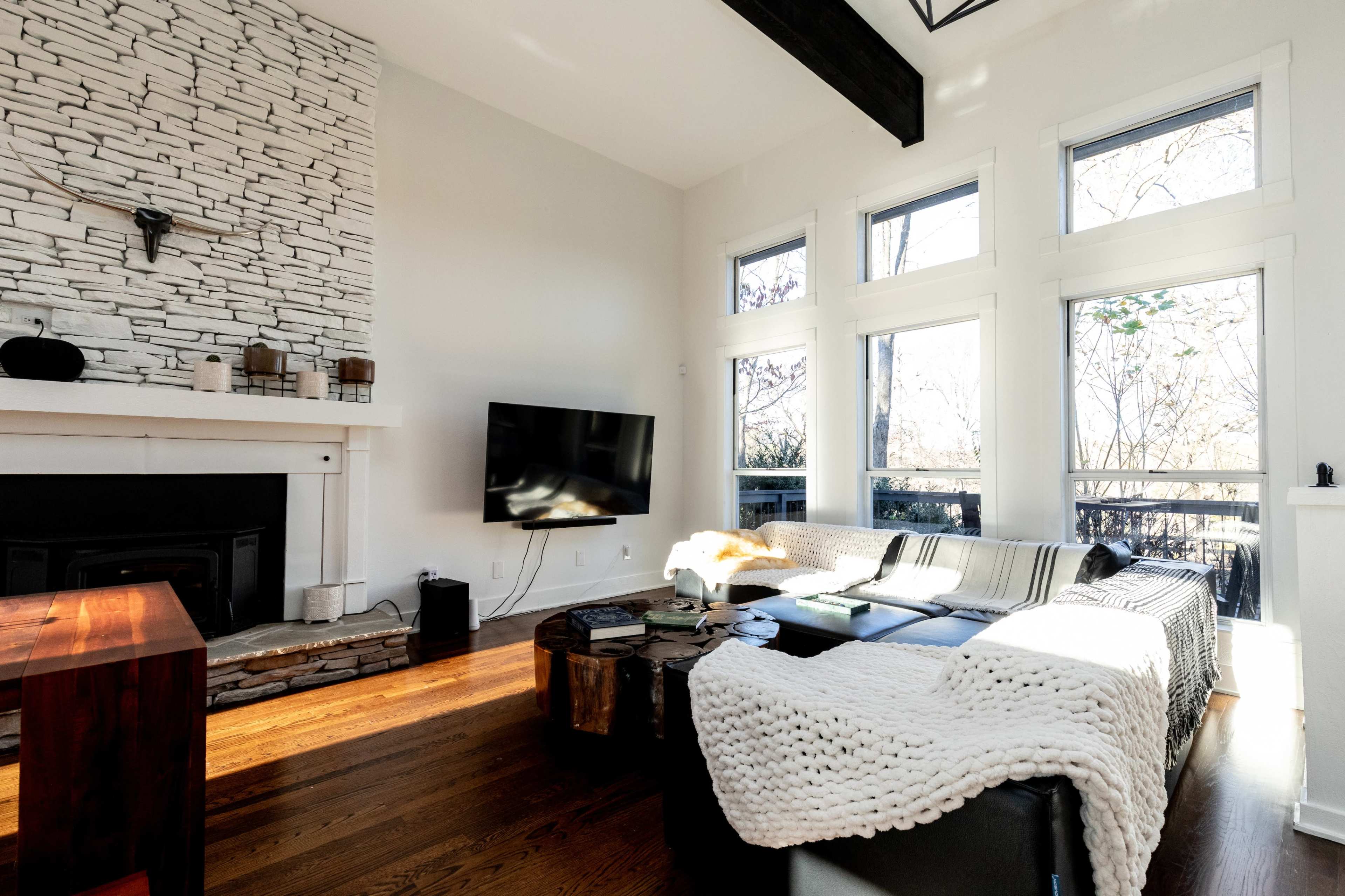 The interior of a well-lit living room features a stone fireplace, a flat-screen TV mounted on the wall, and large windows allowing natural light to fill the space.