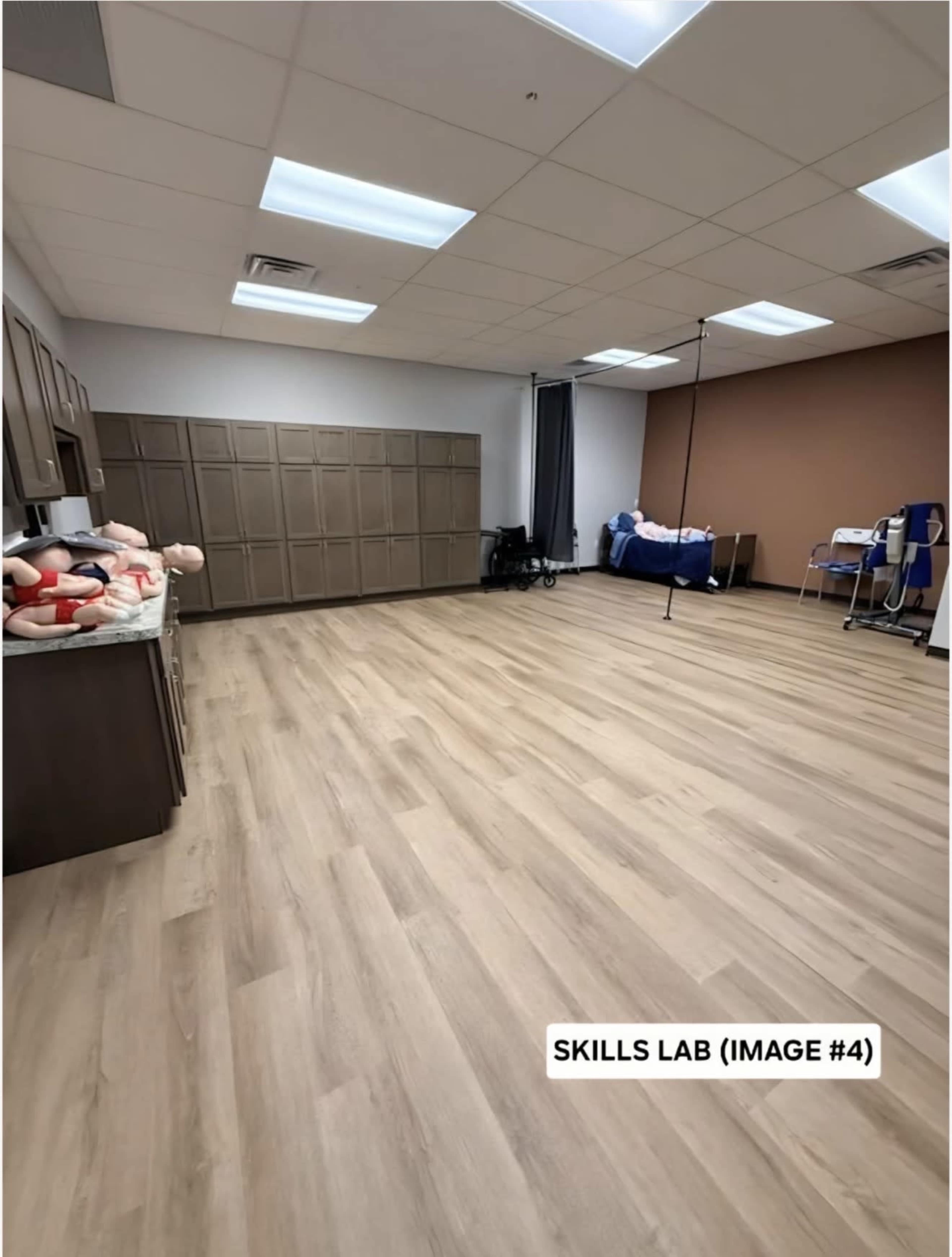 The image shows a spacious skills lab with a long, empty floor, cabinets on one side, a bed in the background, and a few training mannequins positioned near the wall.
