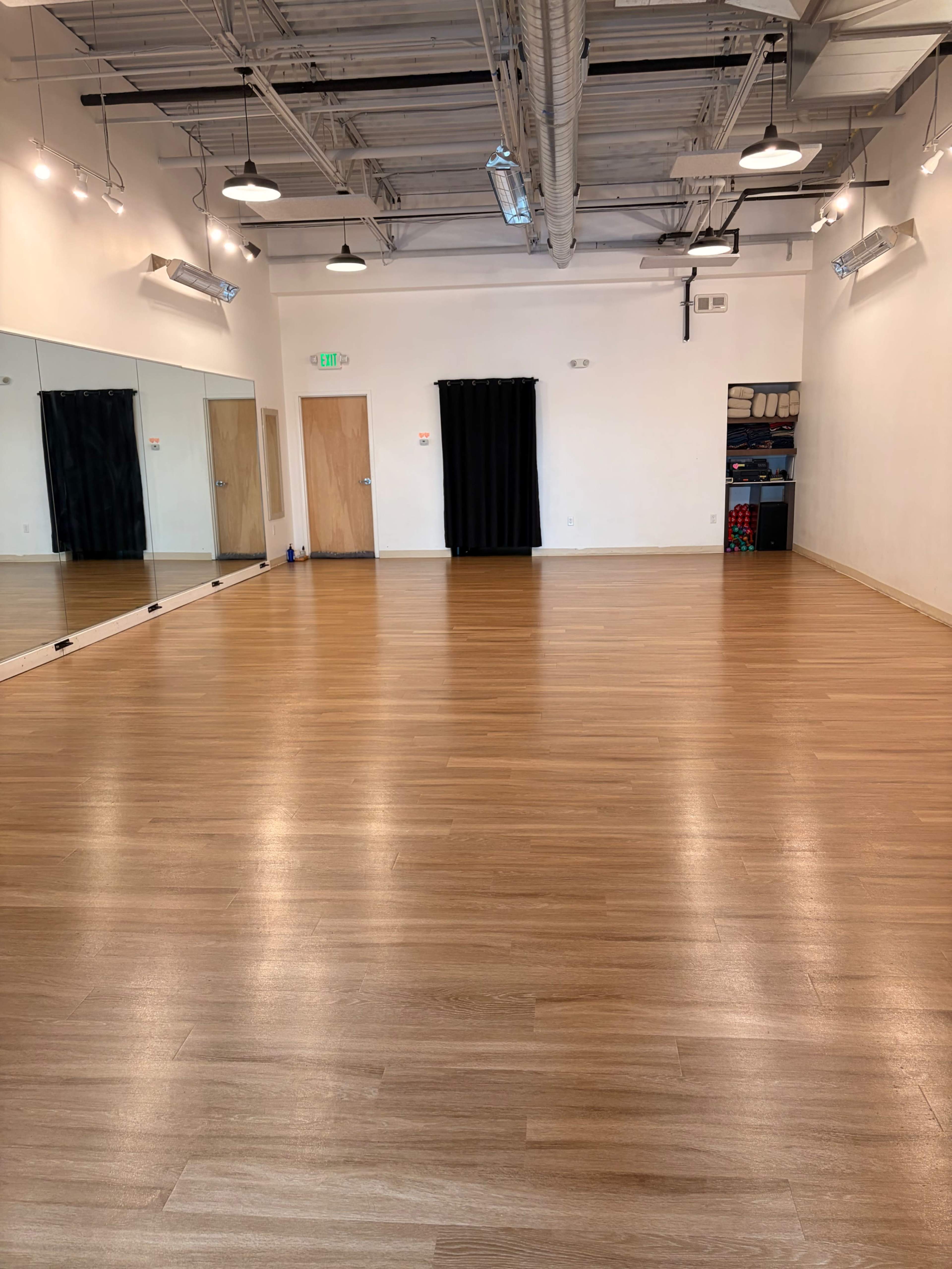 The image shows a spacious, empty room with wooden flooring, a large wall mirror, and a black curtain in the background.